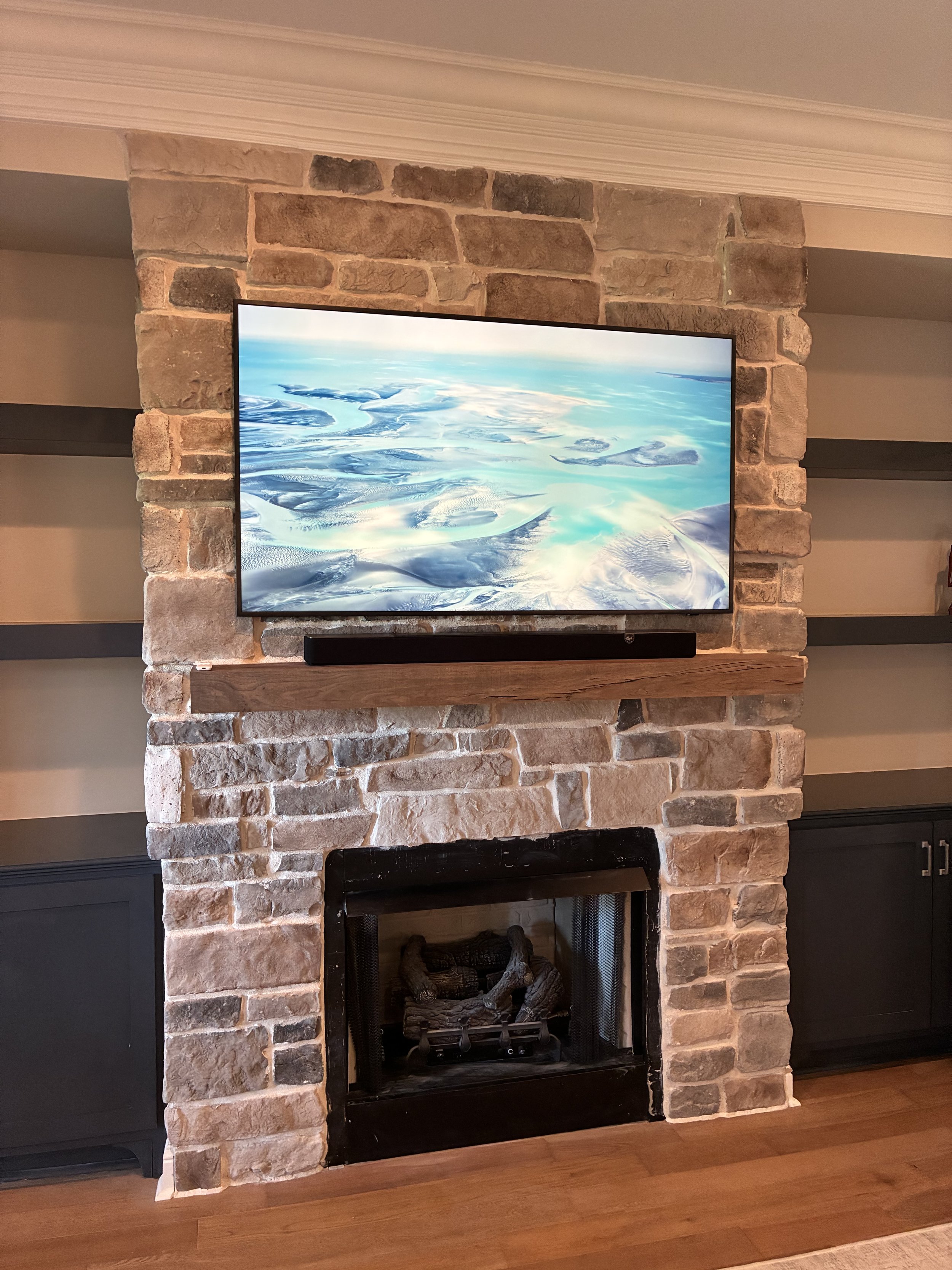 Living room fireplace with a mounted TV above it, stone surround, wooden mantel, and dark cabinets on either side, with a hardwood floor.