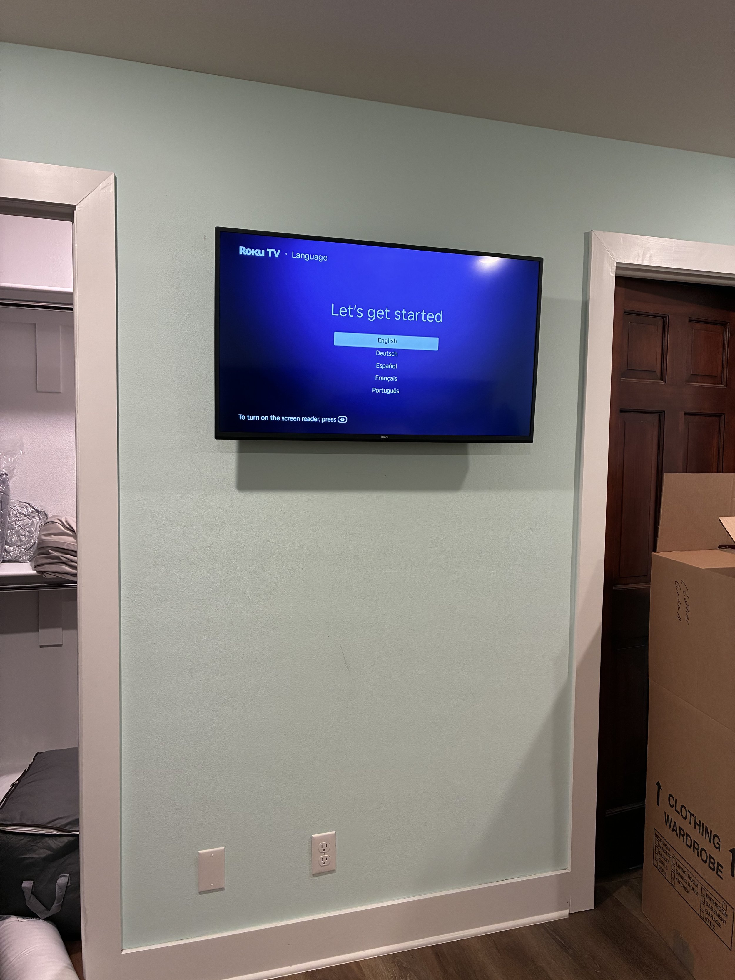 A television mounted on a wall displaying language options for Roku TV. The options include English, Deutsch, Español, Français, and Português.