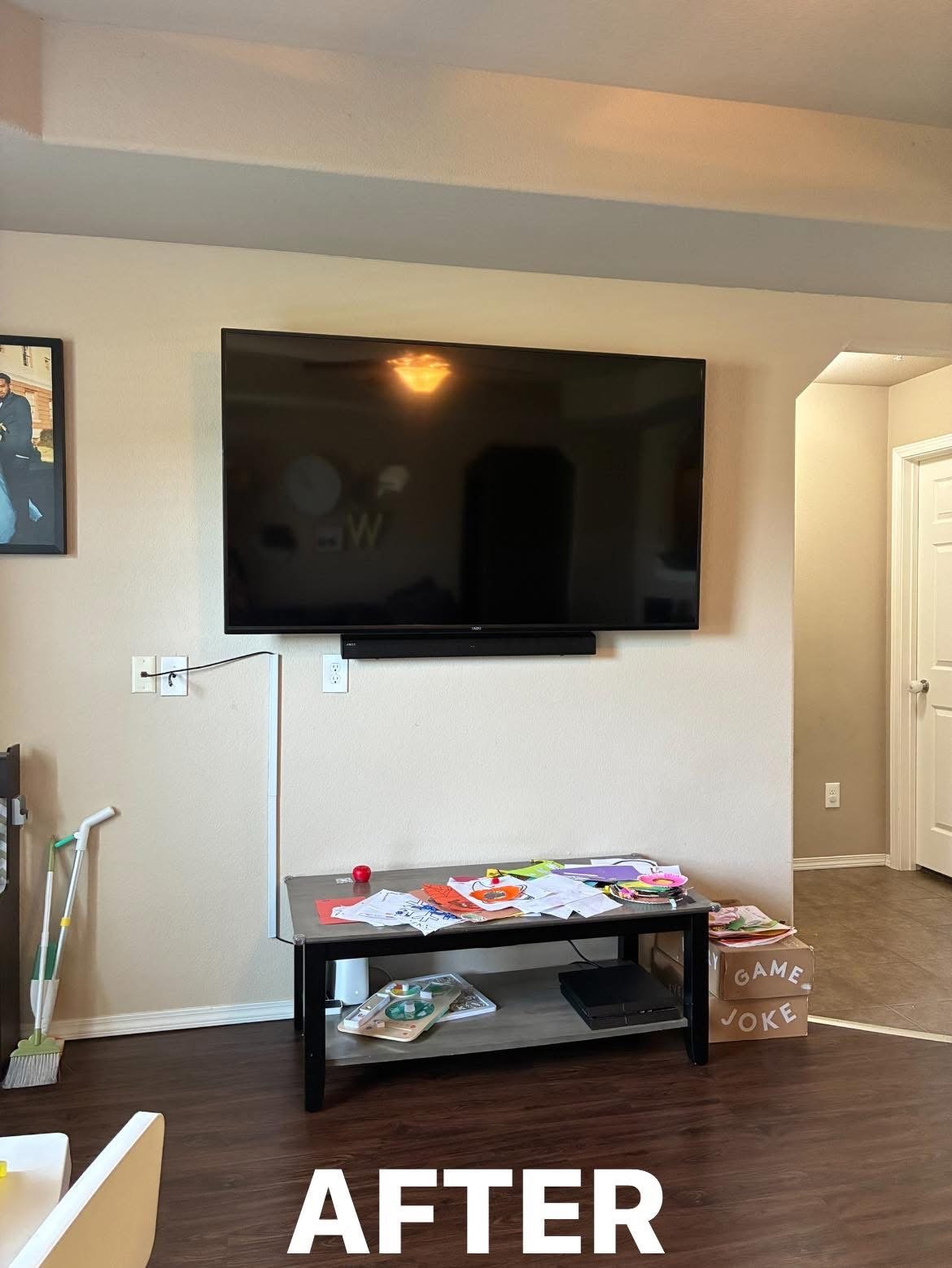 Living room wall with a large flat-screen TV, a black media console below it, a table with scattered papers and some objects, a box labeled 'Game Joke,' and cleaning tools leaning against the wall.