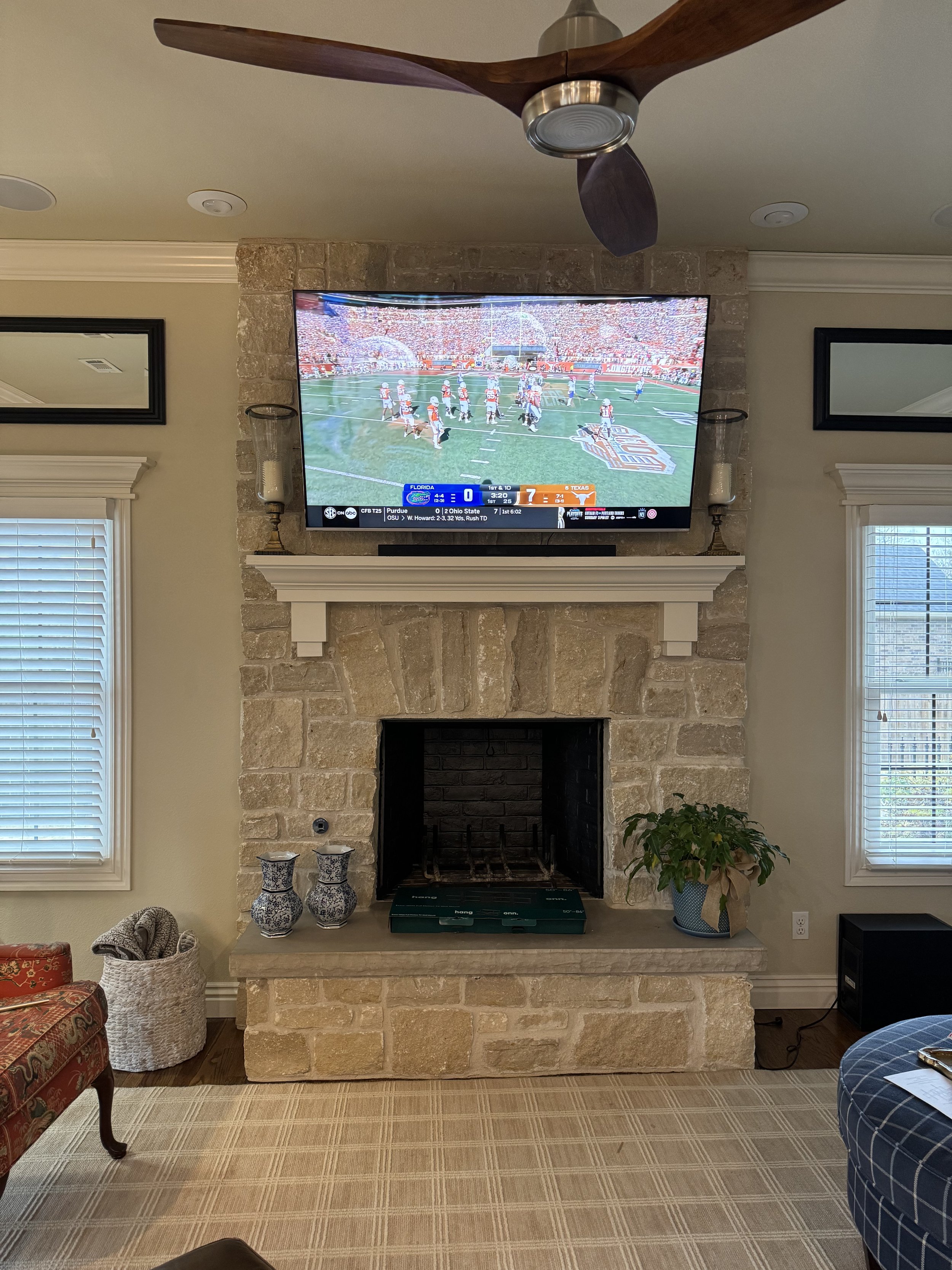 Living room with stone fireplace, mounted flat-screen TV showing a football game, ceiling fan, two windows, decorative vases, a potted plant, and furniture.