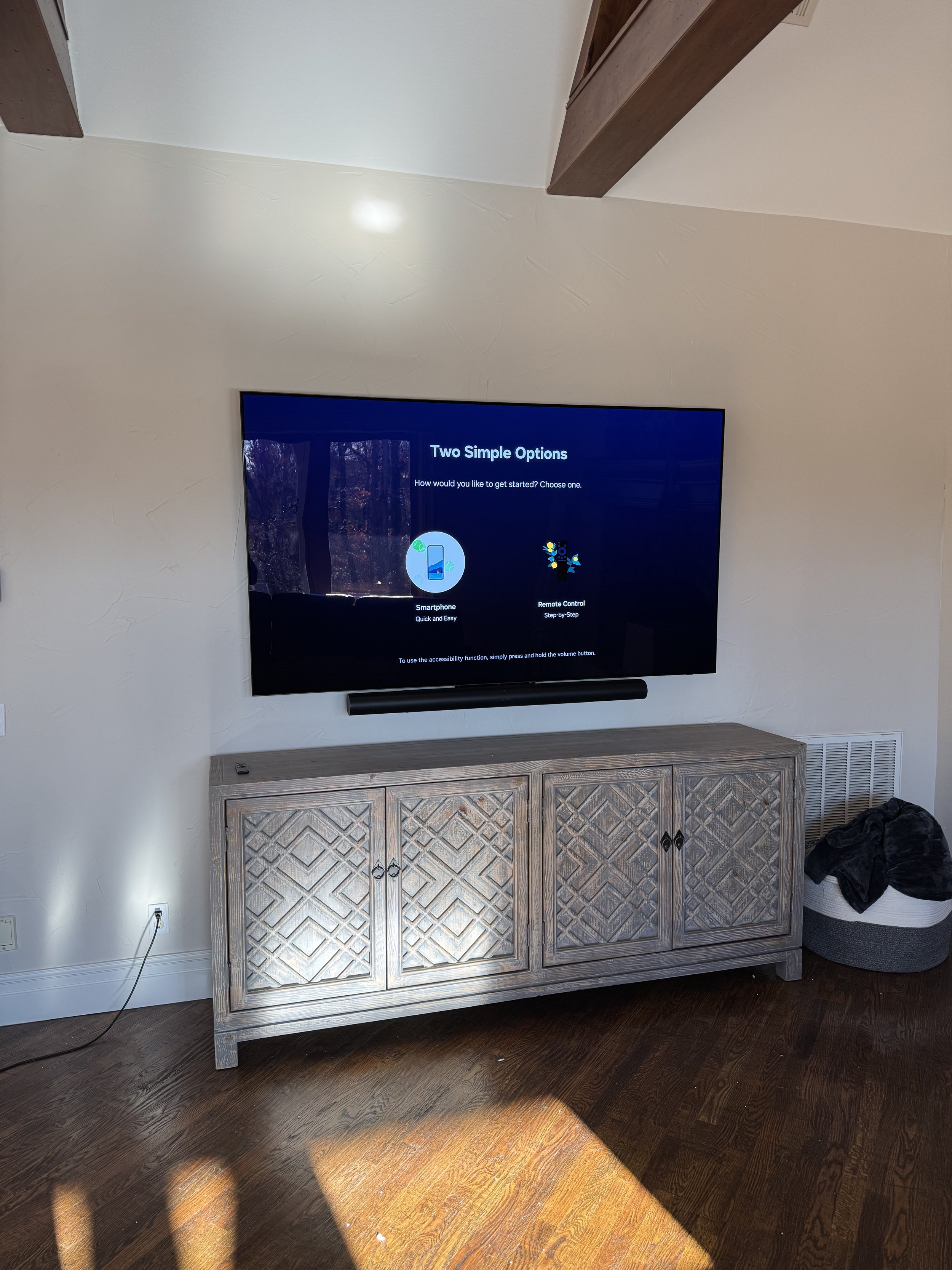 Living room with a large flat-screen TV mounted on the wall, displaying setup options, above a wooden cabinet. Sunlight on hardwood floor, with a black and white round storage basket and clothing on the right side.