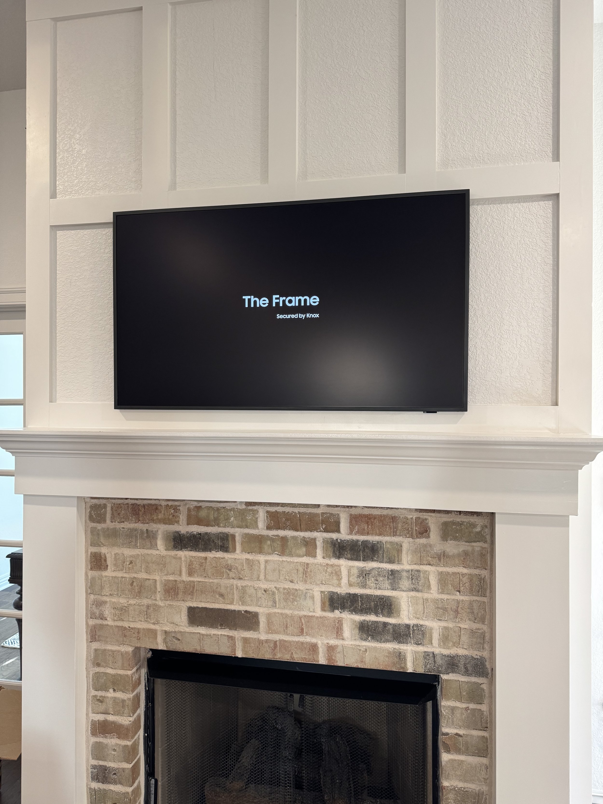 Flat-screen TV mounted above a brick fireplace with a white wood mantel surround, in a living room.