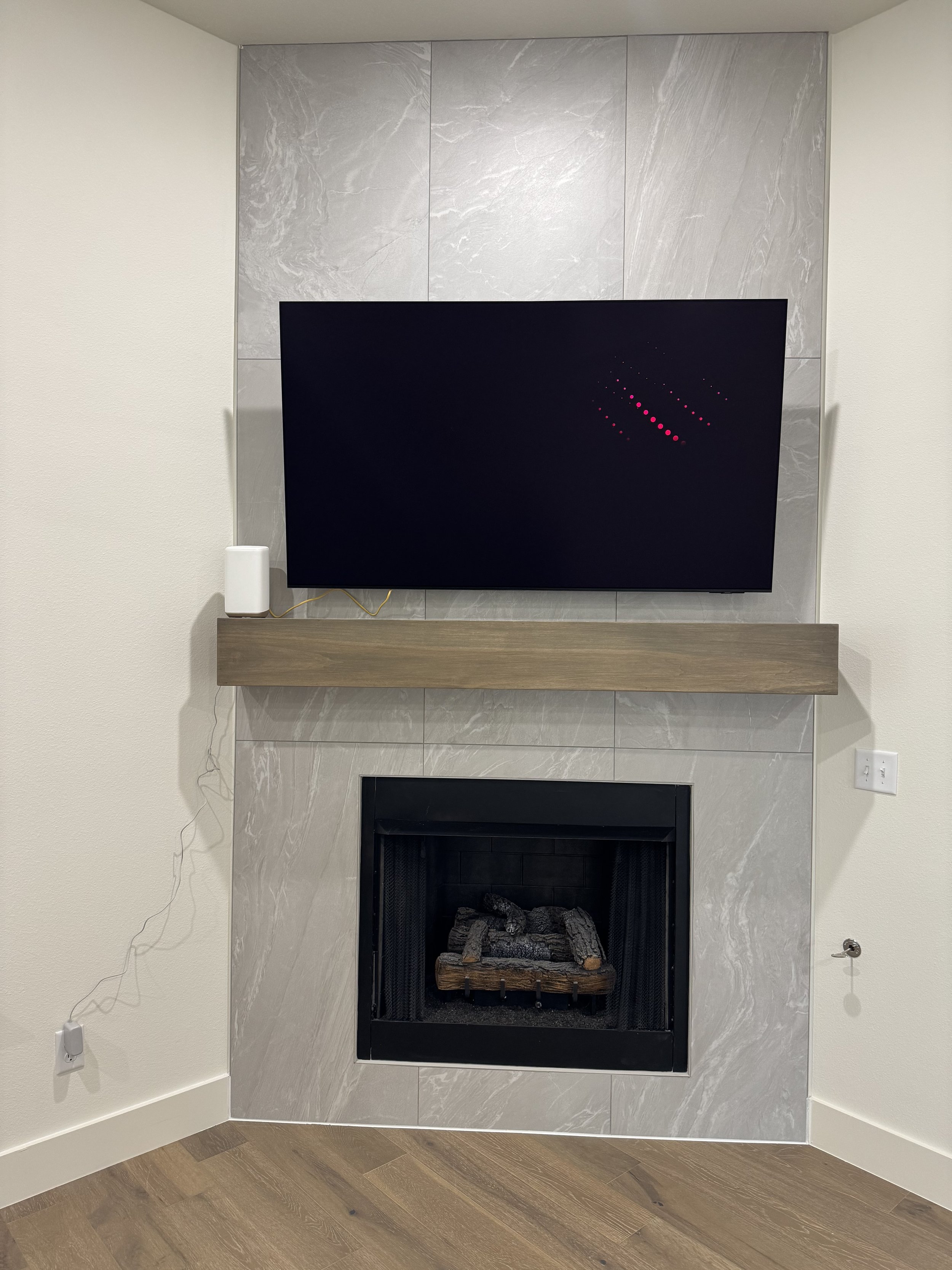 Living room corner with a modern fireplace, gray marble surround, and a wall-mounted flat-screen TV above it. A small white speaker is to the left of the TV on a wooden mantel.