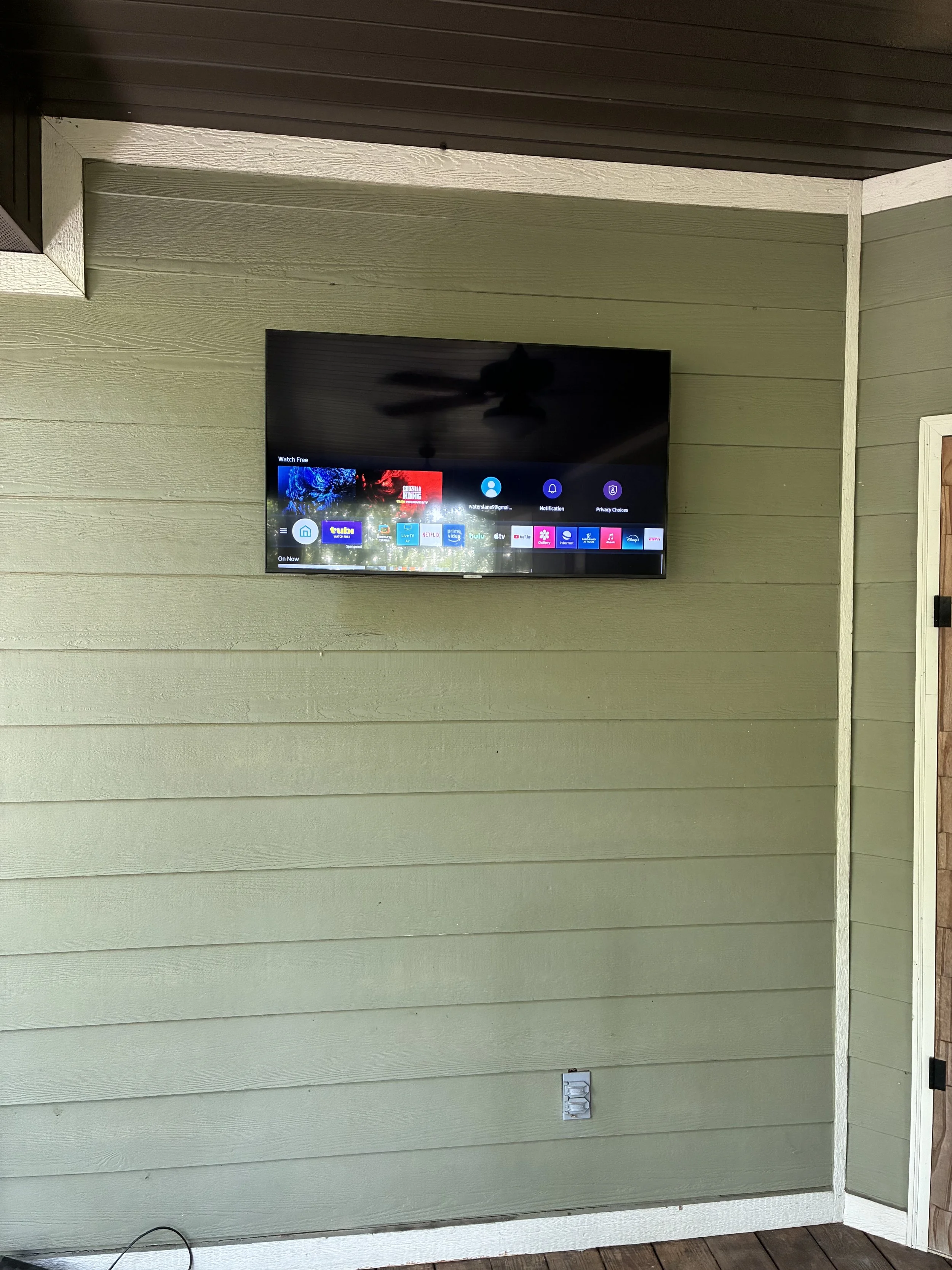 A flat-screen TV mounted on a green wooden wall outdoors, displaying a streaming service menu. An electrical outlet and a black cable are visible near the floor.