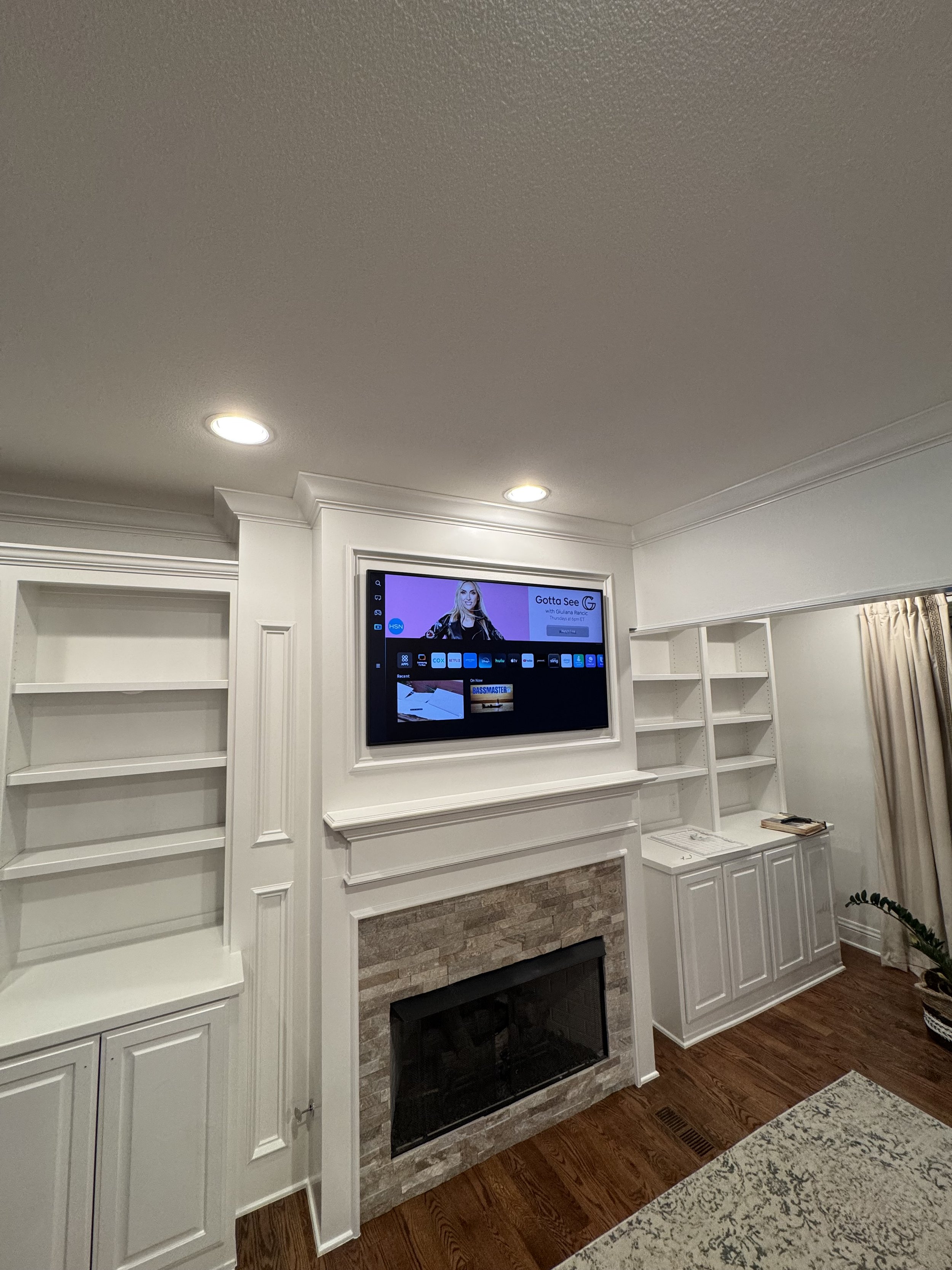 Living room with built-in white shelves on either side of a fireplace, with a flat-screen TV mounted above the fireplace, and hardwood floors.