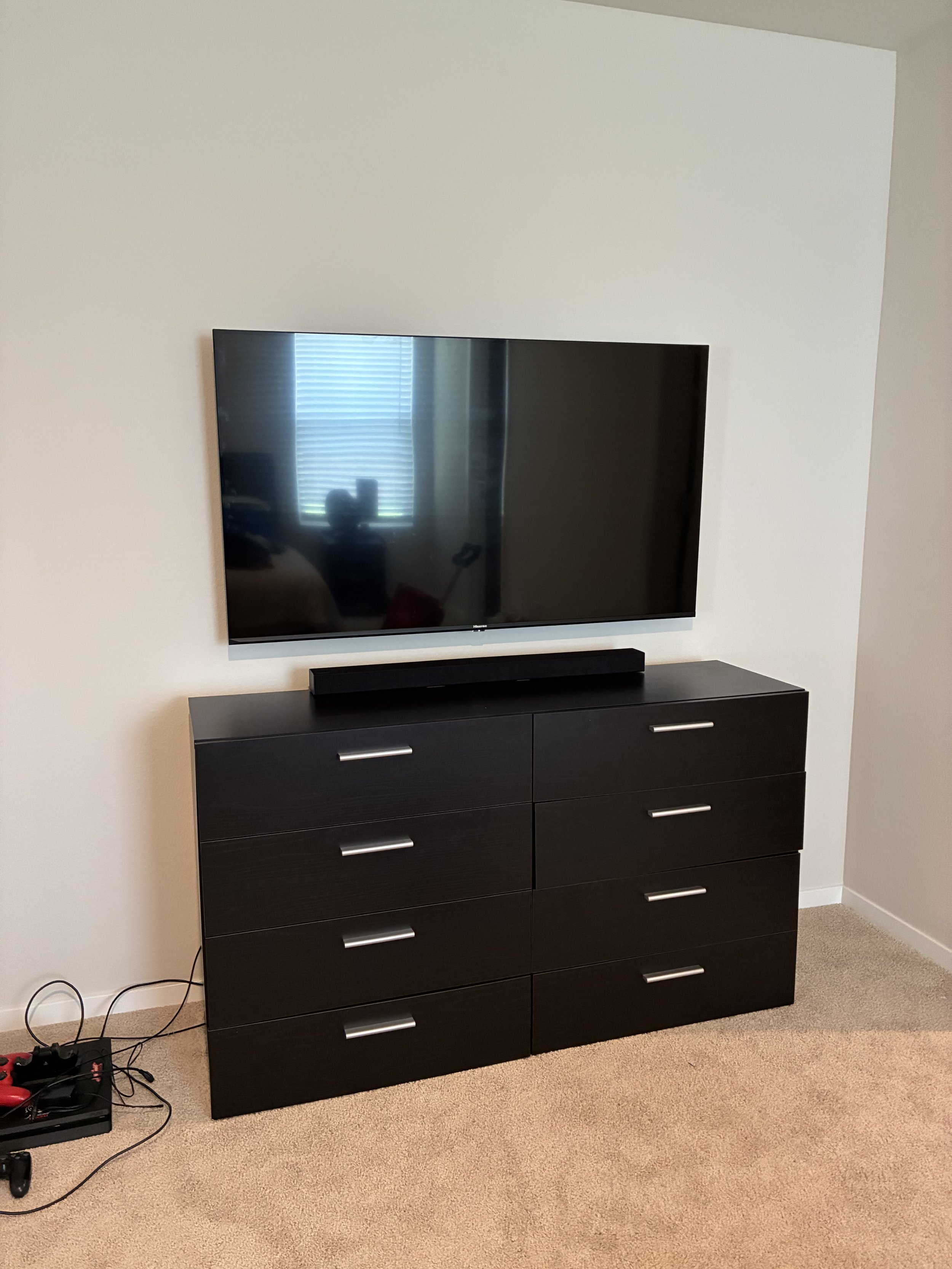 A flat-screen TV mounted on a white wall above a black dresser with silver handles, sitting on a beige carpet. A soundbar is placed on top of the dresser, and a gaming console with controllers is on a small black stand on the floor nearby.