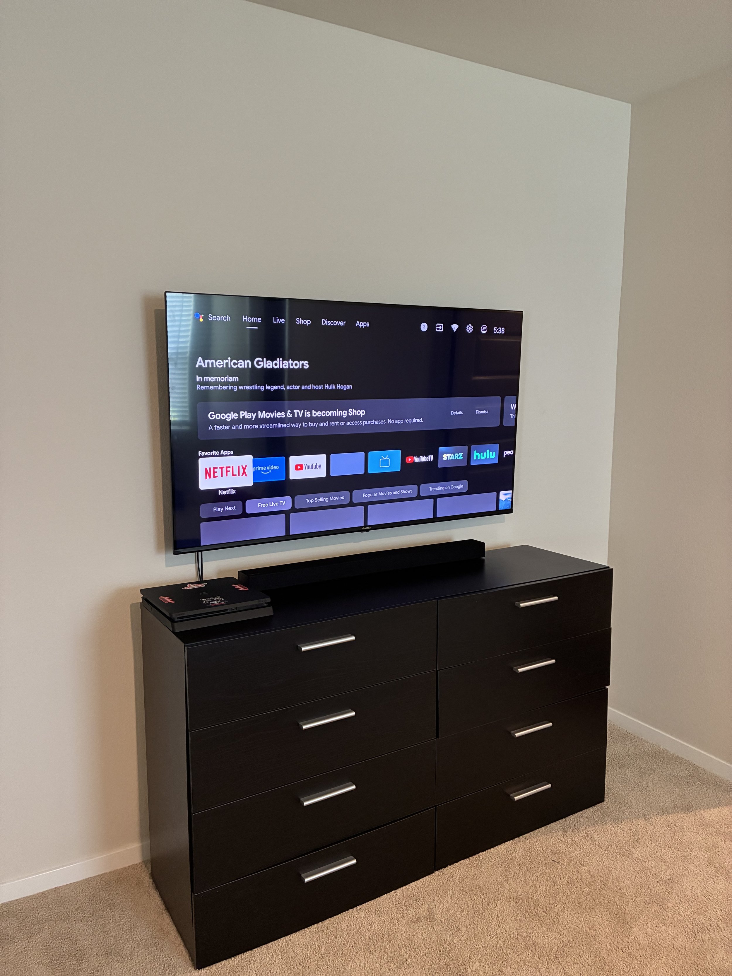 A flat-screen TV mounted on a wall above a black dresser with six drawers in a room with beige carpet and off-white walls.