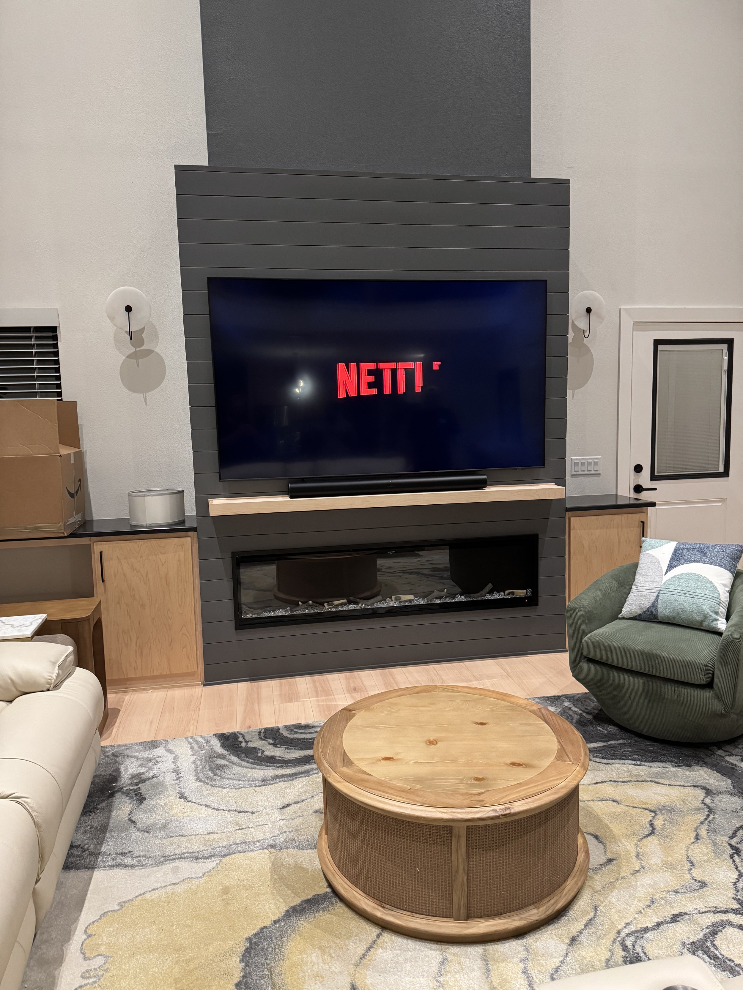 Living room with a mounted flat-screen TV displaying the Netflix logo, a fireplace beneath the TV, and seating furniture including a beige sofa and a green armchair with a pillow. There is a round wooden coffee table in the center, a patterned rug, w