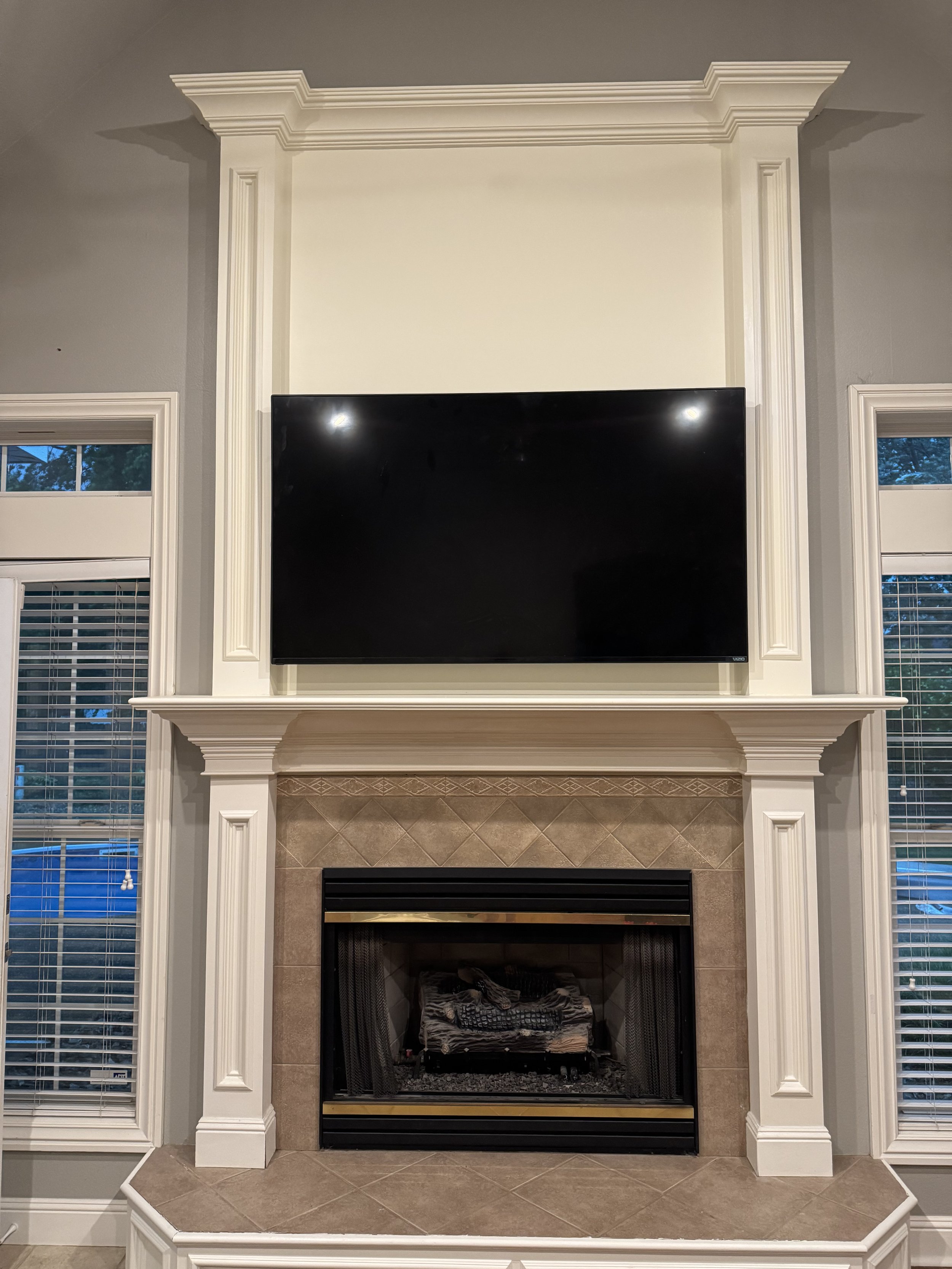 Living room fireplace with a mounted flat-screen TV above, flanked by two windows with white blinds, decorative molding around the fireplace, and a granite hearth.