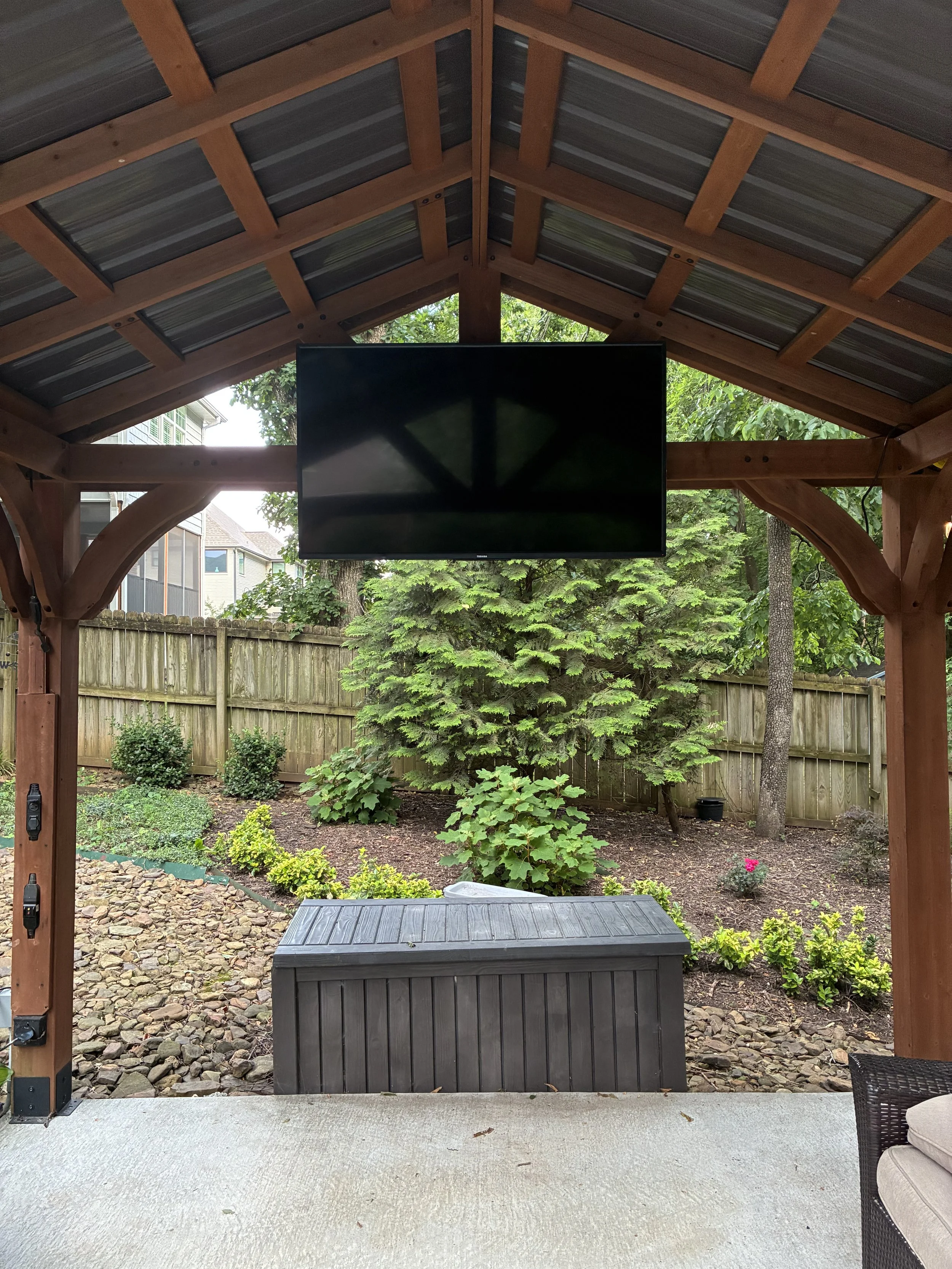 Outdoor covered patio with a mounted flat-screen TV, a storage box, a small section of cushioned seating, and a landscaped yard with trees, shrubs, and a wooden fence.