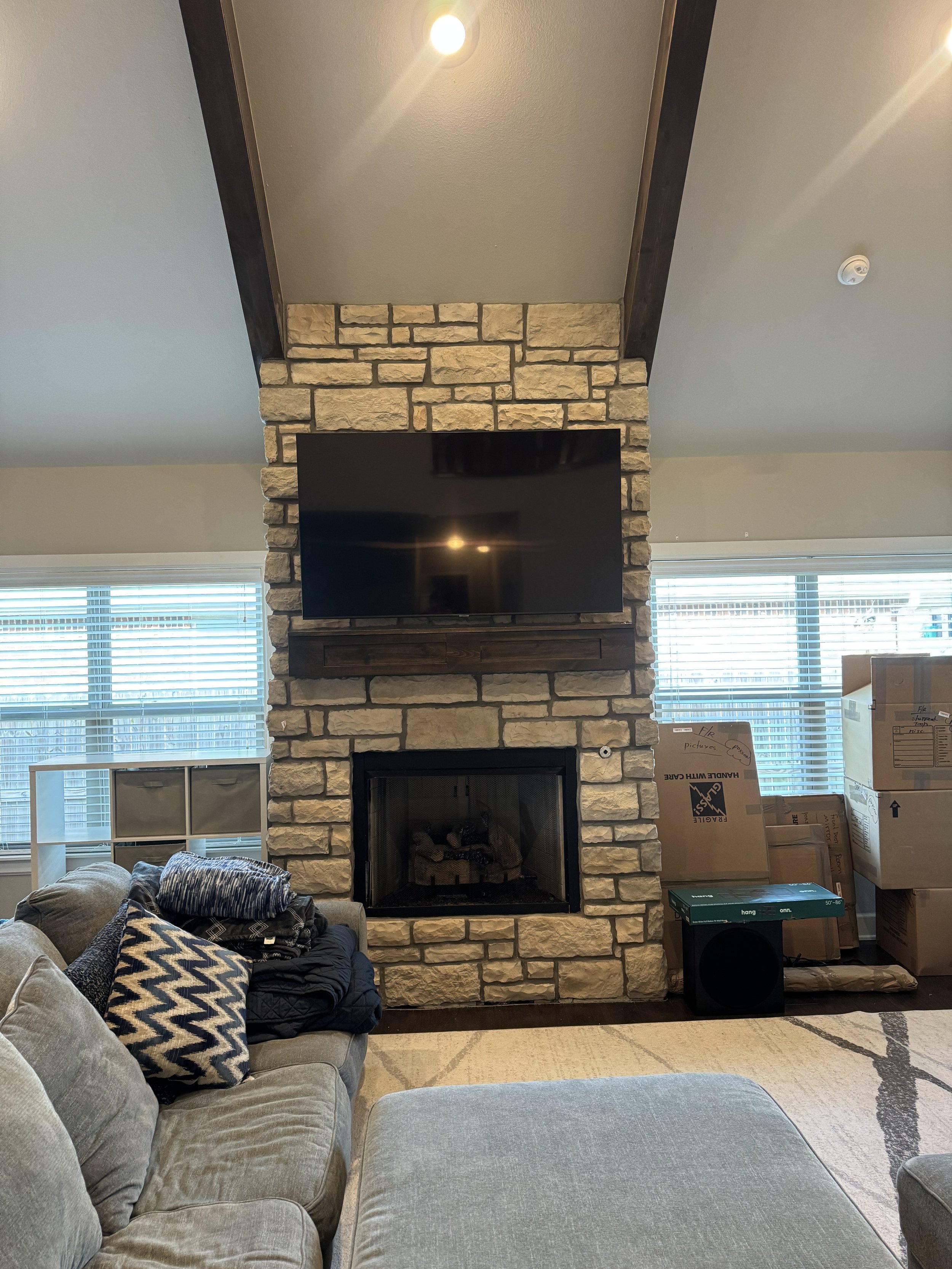 Living room with a stone fireplace, a mounted television above the fireplace, and a gray couch with pillows and jackets nearby. There are boxes and a speaker on the right side.