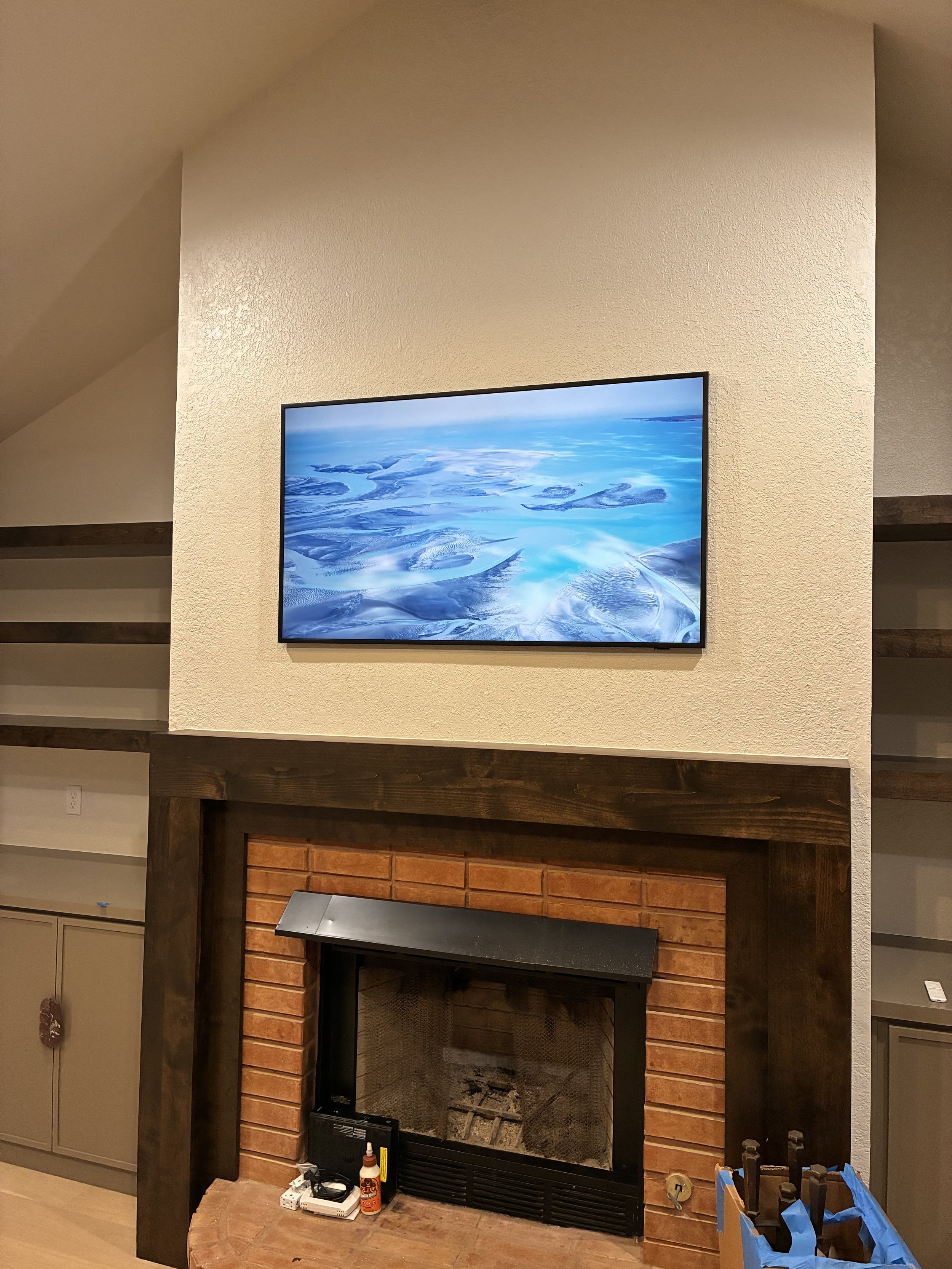 A living room fireplace with a brick surround and a black metal hood, with a flat-screen TV mounted above it showing a blue landscape image. There are built-in shelves and cabinets on either side of the fireplace.