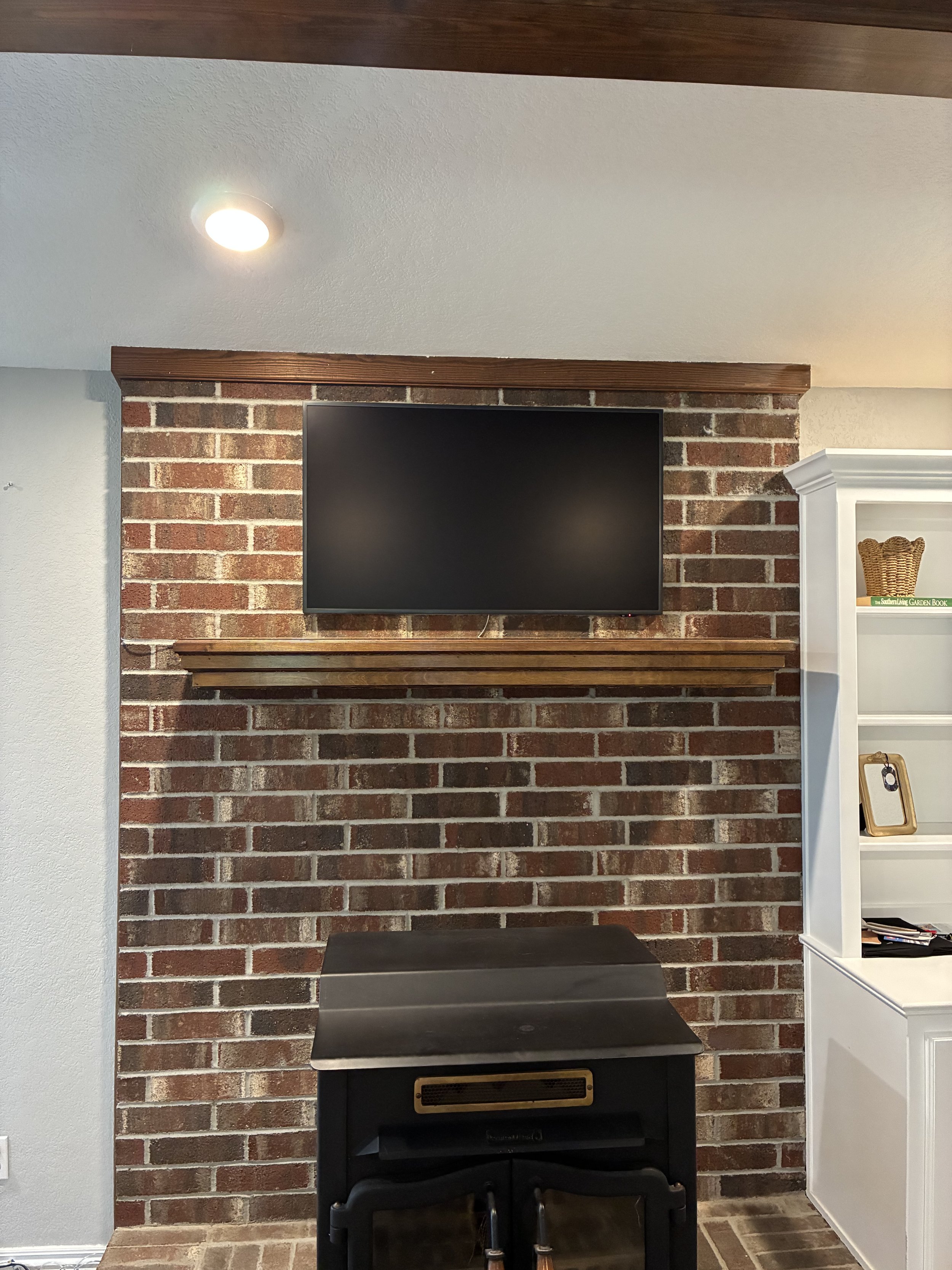 Living room with brick wall, mounted flat-screen TV, black wood stove, white shelving unit with books and decorative items.
