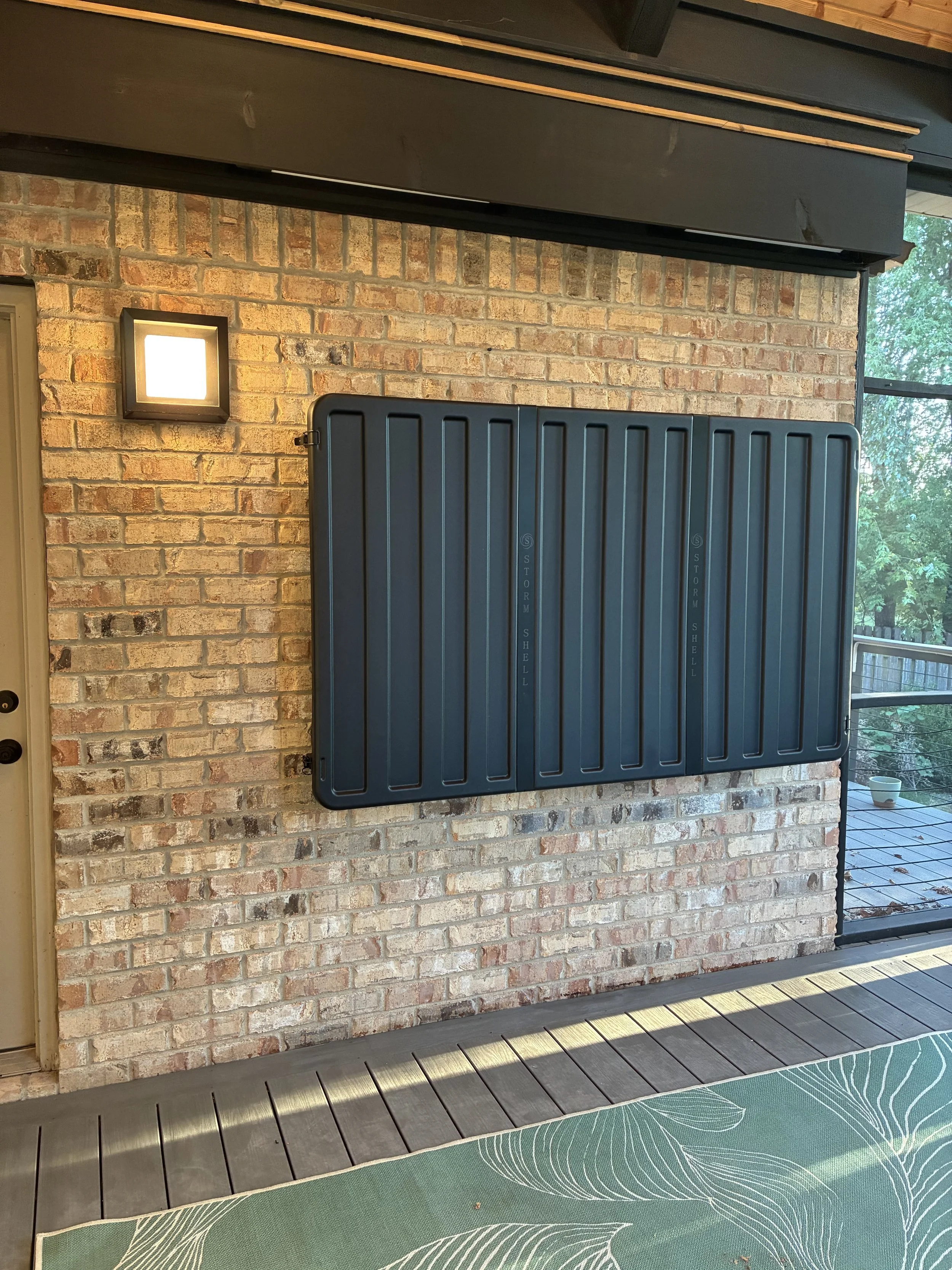 A black Storm Shell patio cover folded against a brick wall, next to a square wall light and a sliding glass door leading to a deck with a potted plant.