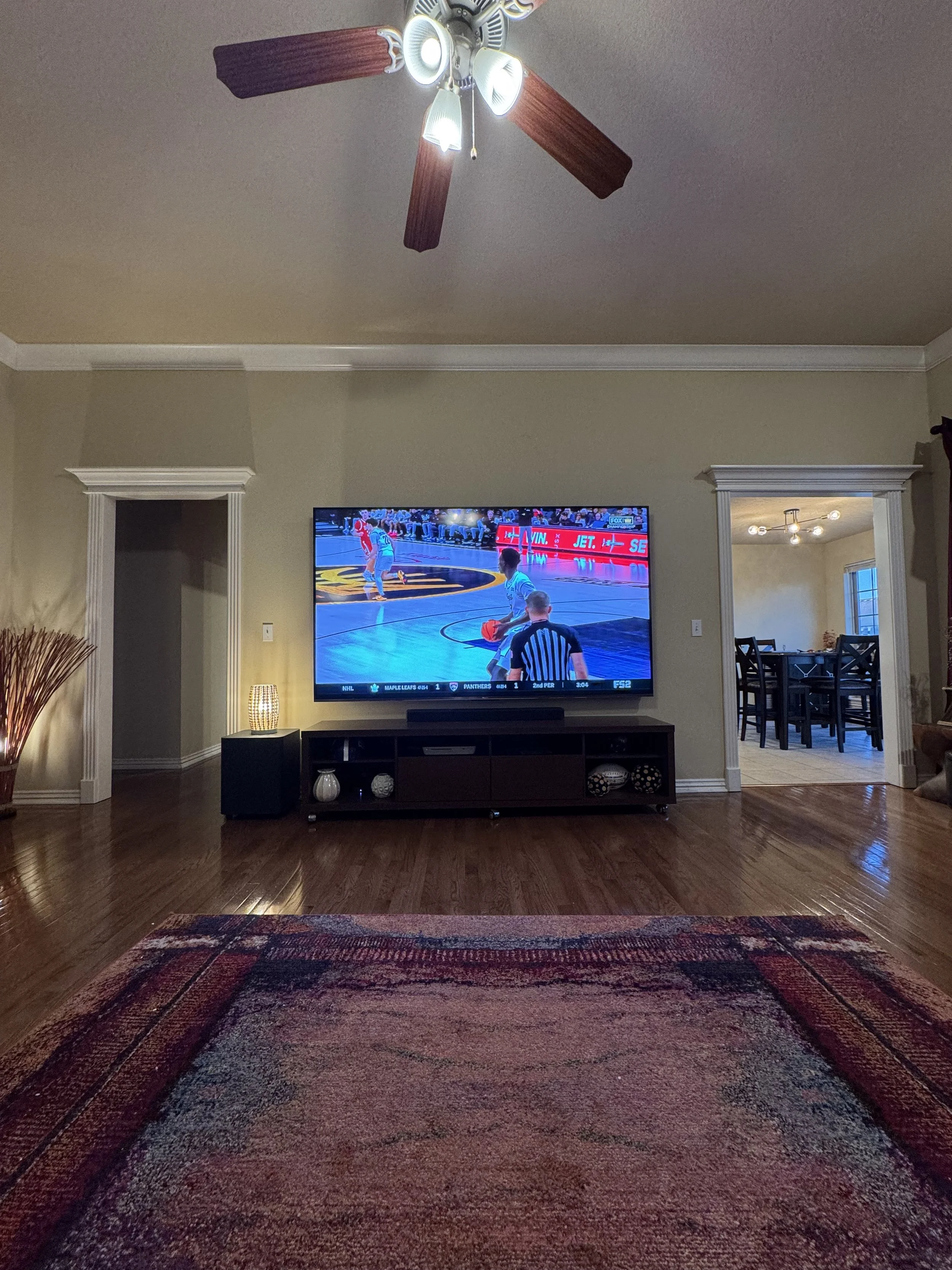 Living room with a large flat-screen TV showing a basketball game, a wooden floor, a rug, a ceiling fan with lights, a small speaker and decorative items on a shelf, and a dining room visible through a doorway.