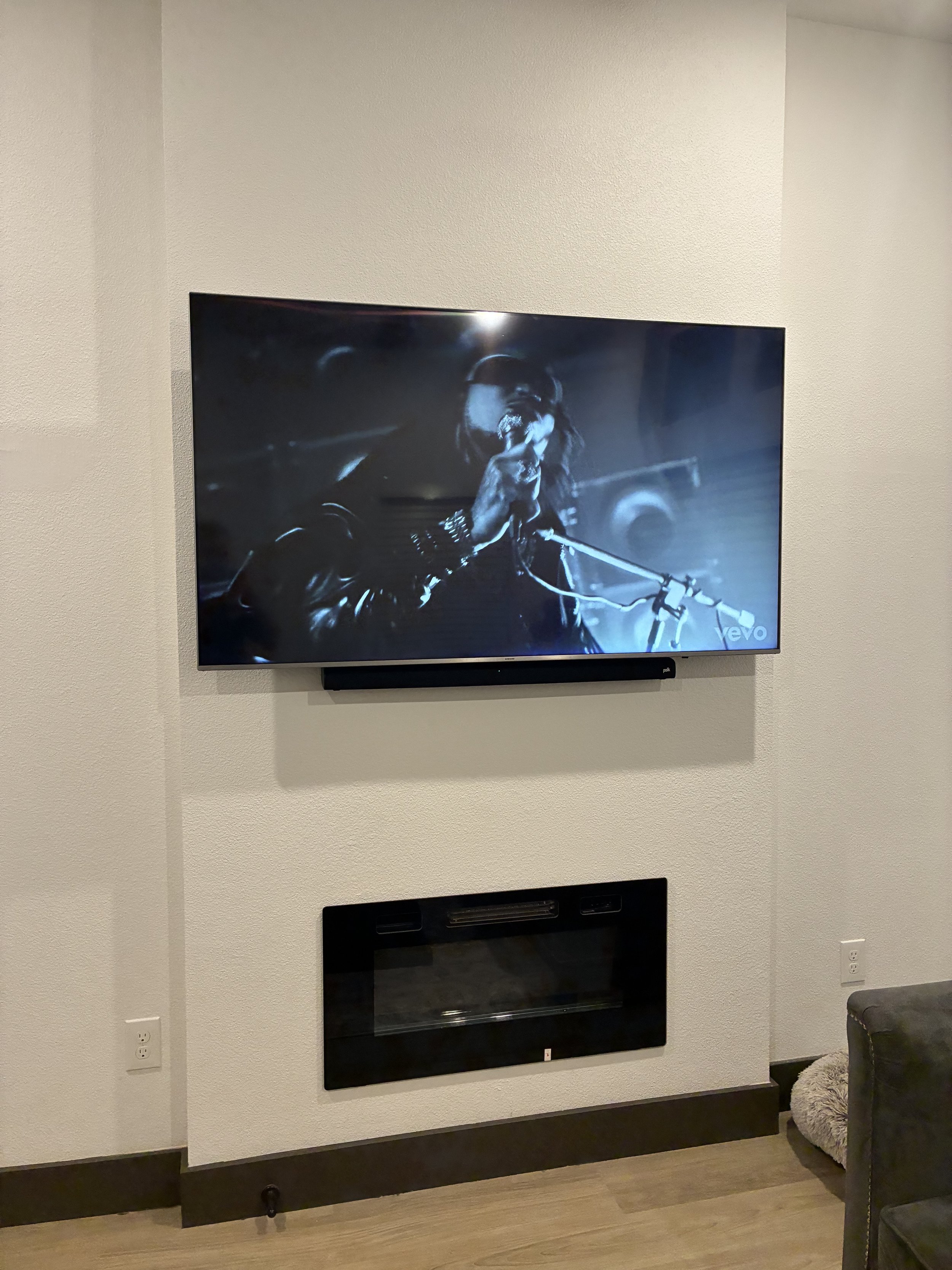 A television mounted on a wall displaying a black and white image of a person singing into a microphone. Below the TV is a fireplace with a glass front, and to the right is part of a gray chair and a cat bed on the floor.