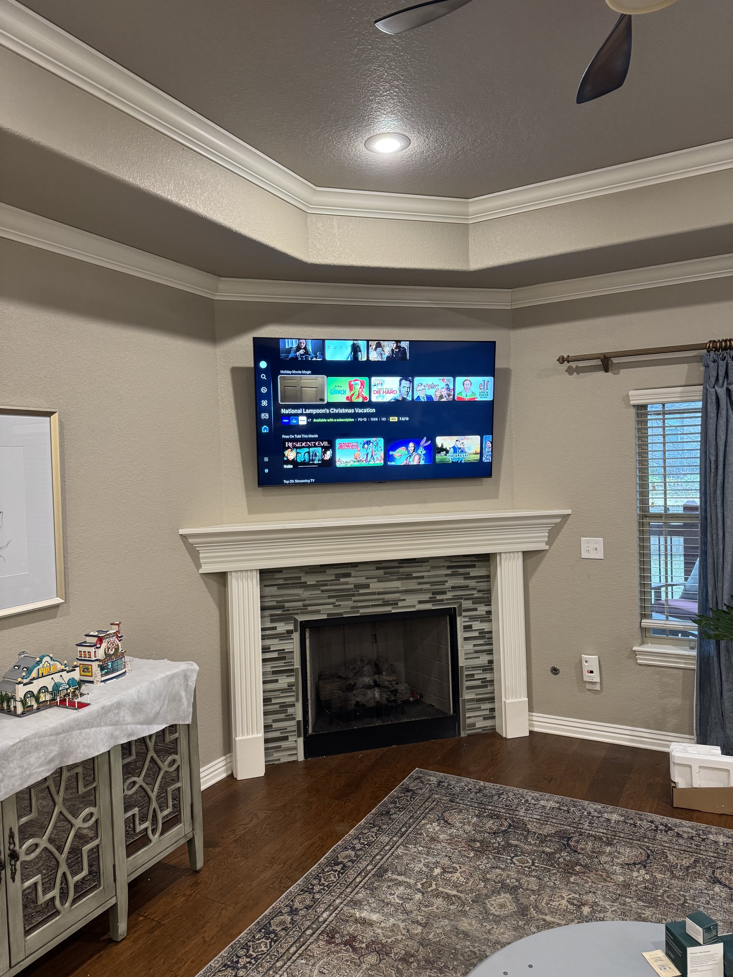 Living room with a flat-screen TV mounted above a fireplace, a window with blue curtains, a rug, and a table with model buildings.