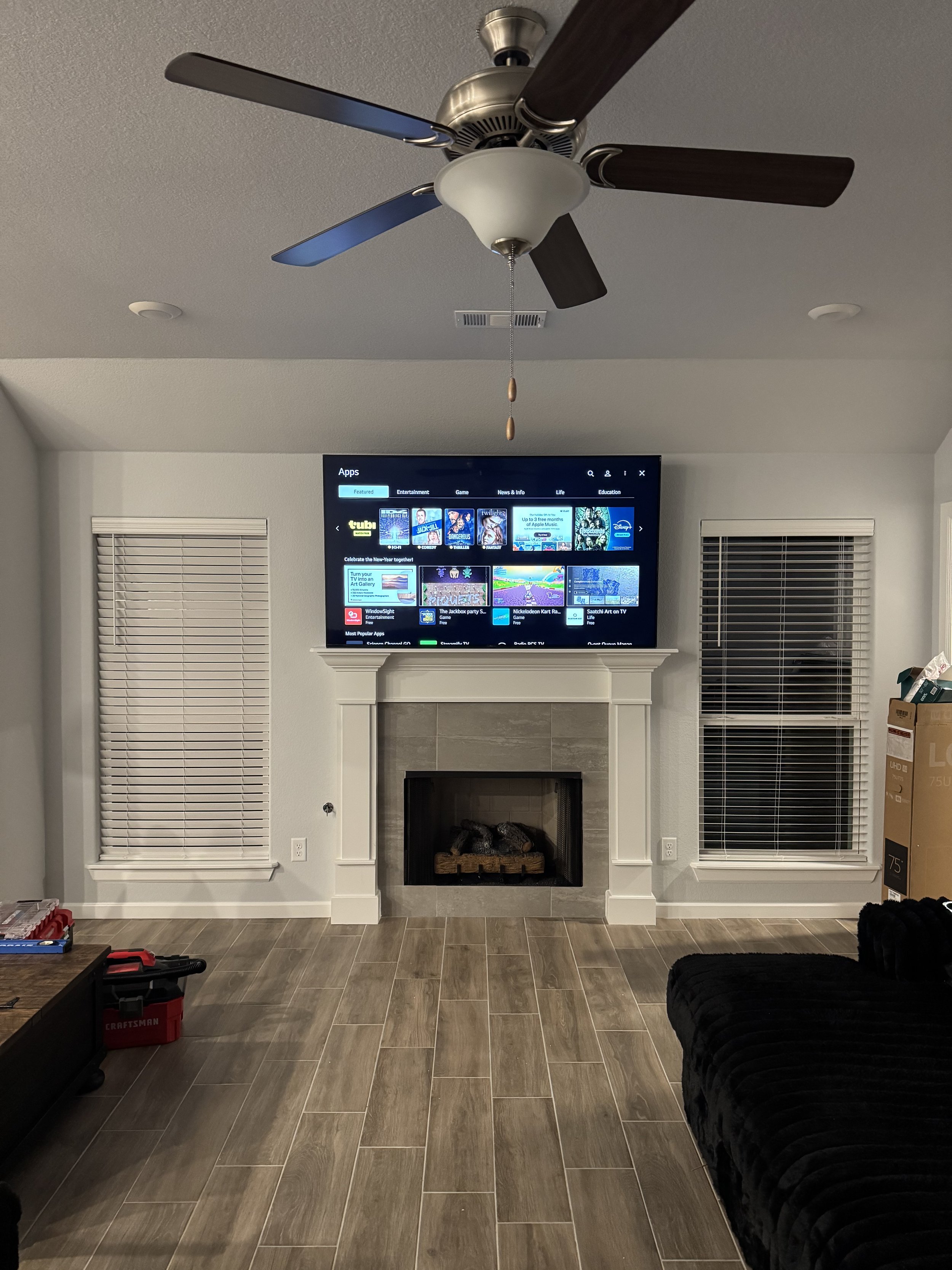 Living room with a ceiling fan, mounted flat-screen TV above a fireplace, and two windows with blinds. There is a black couch and some tools or boxes in the room.