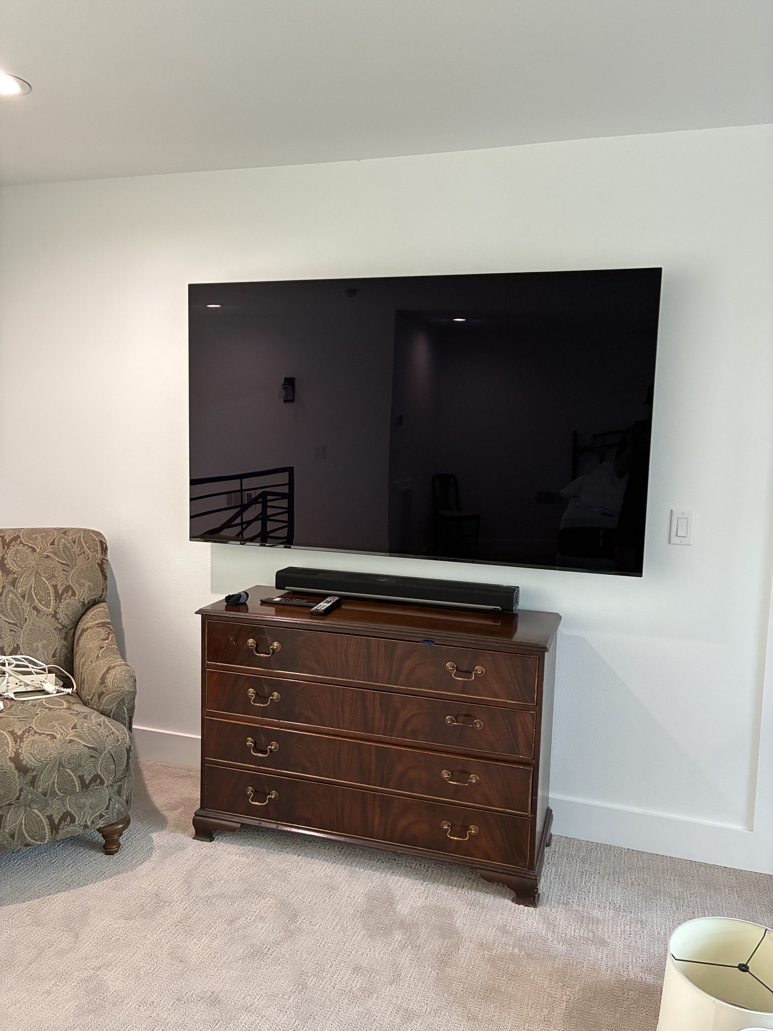 Living room with a large flat-screen TV mounted on a white wall, a wooden dresser beneath it, and an upholstered armchair nearby.