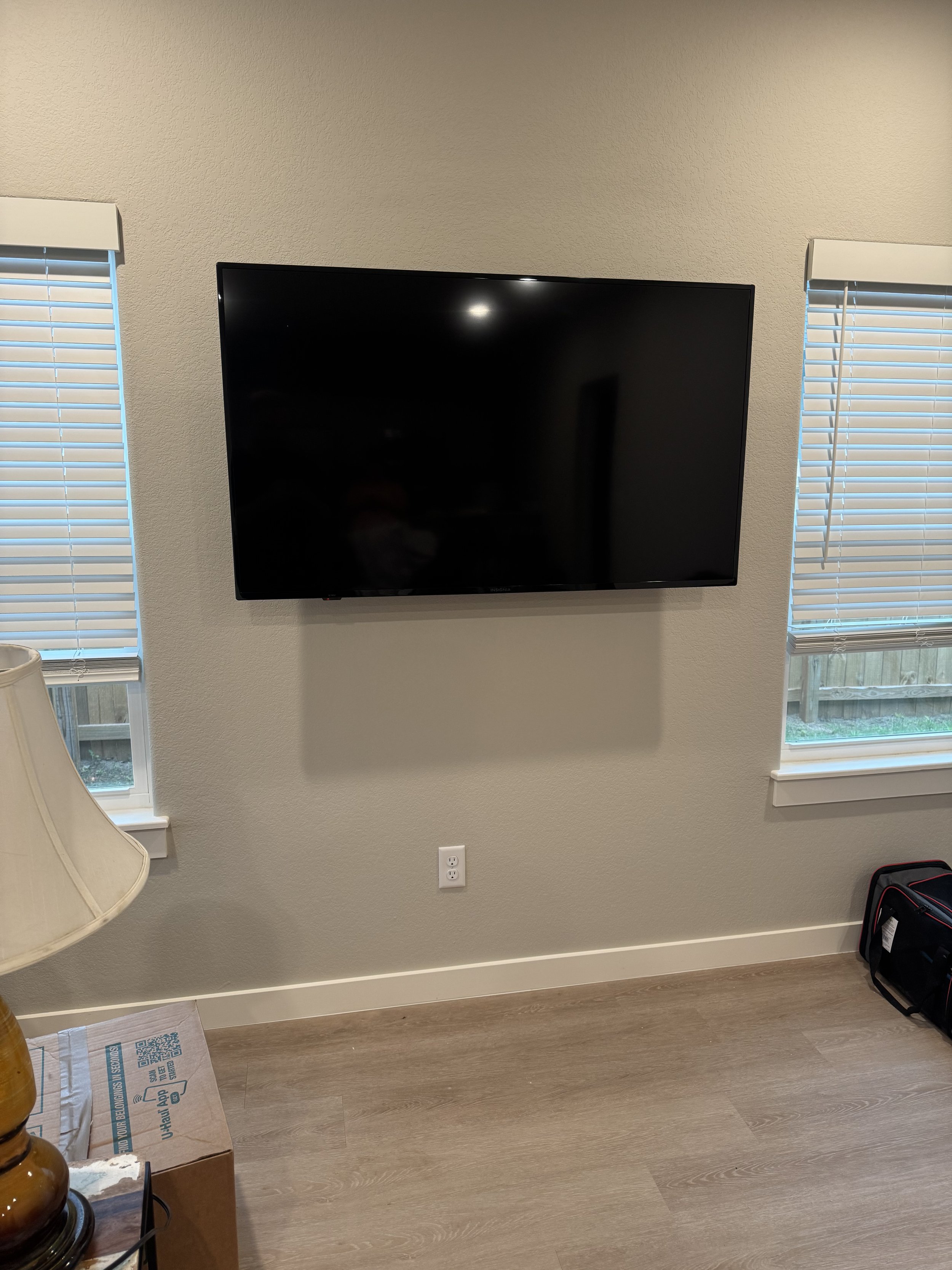 A flat-screen TV mounted on a beige wall between two windows with closed blinds, with a power outlet below and a backpack on the floor to the right. A lamp and a cardboard box are partially visible on the left.