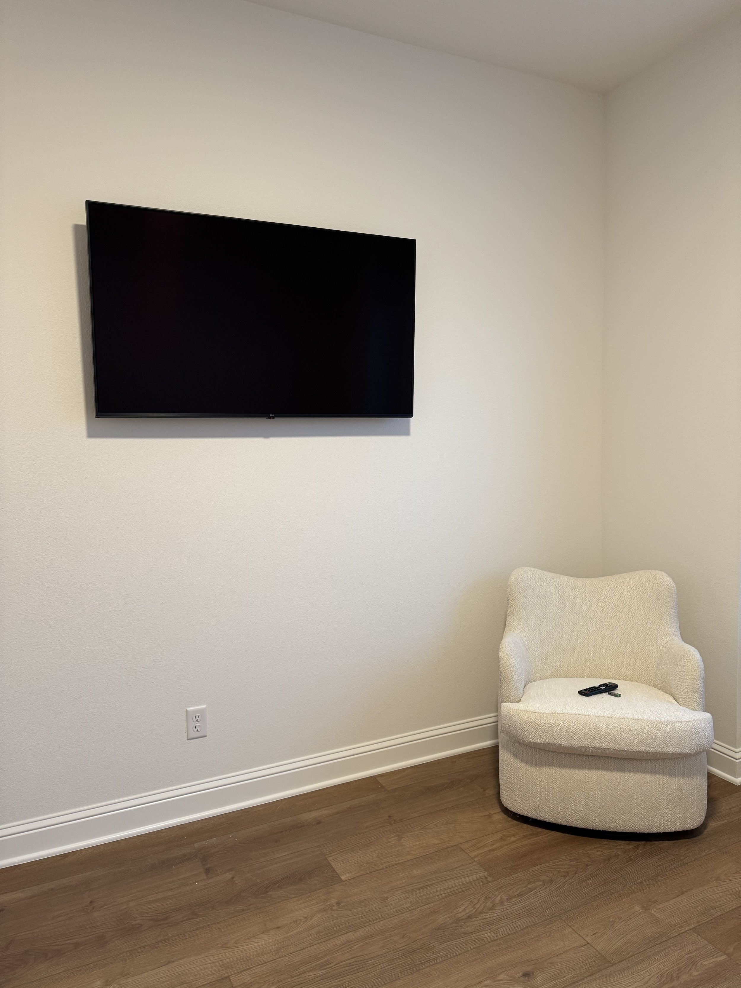 A TV mounted on a white wall with a beige armchair below it and a remote control on the chair.