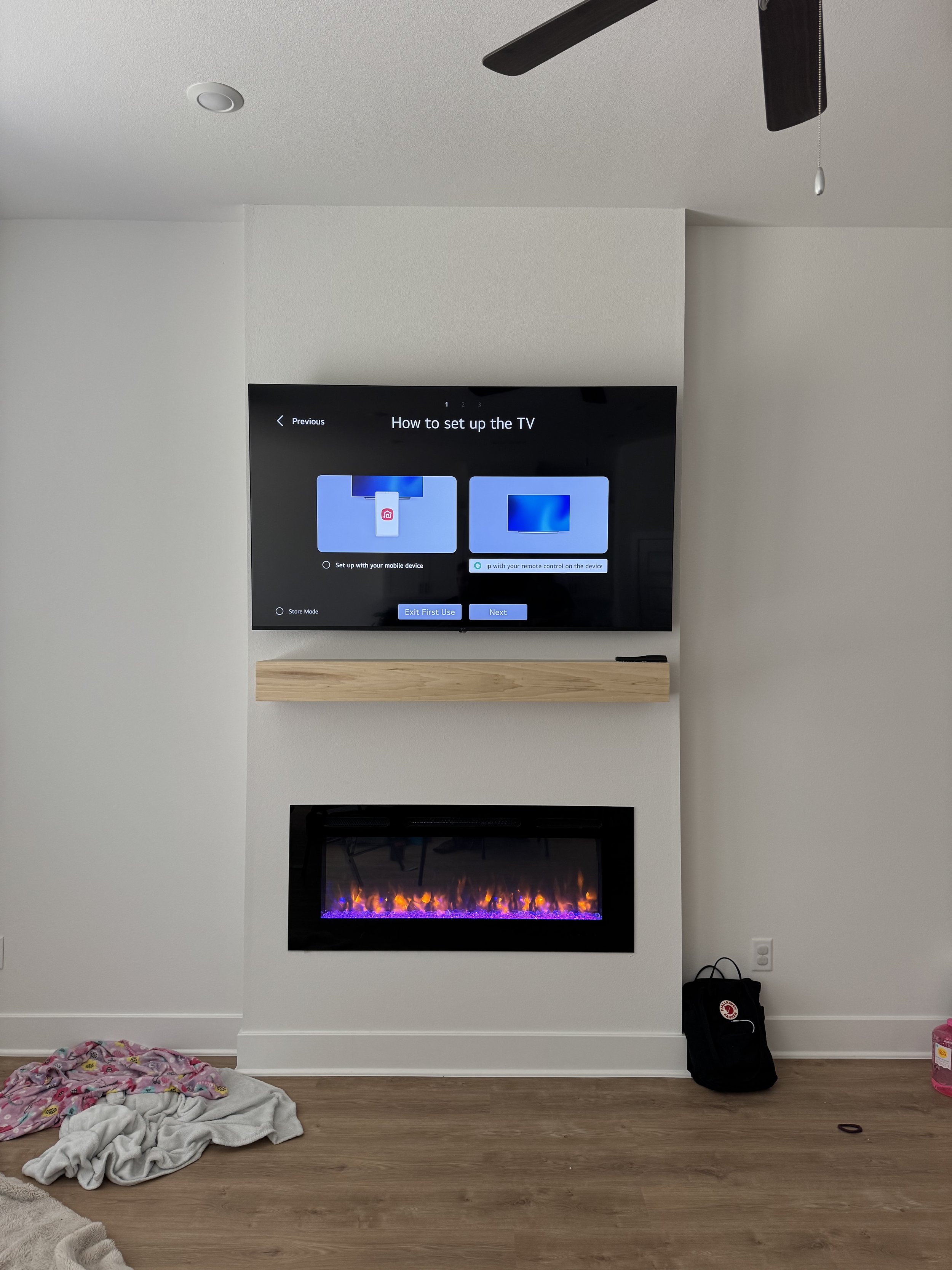 Living room with wall-mounted TV displaying setup instructions, electric fireplace below, ceiling fan above with pull chain, some clothes and a bag on the floor.