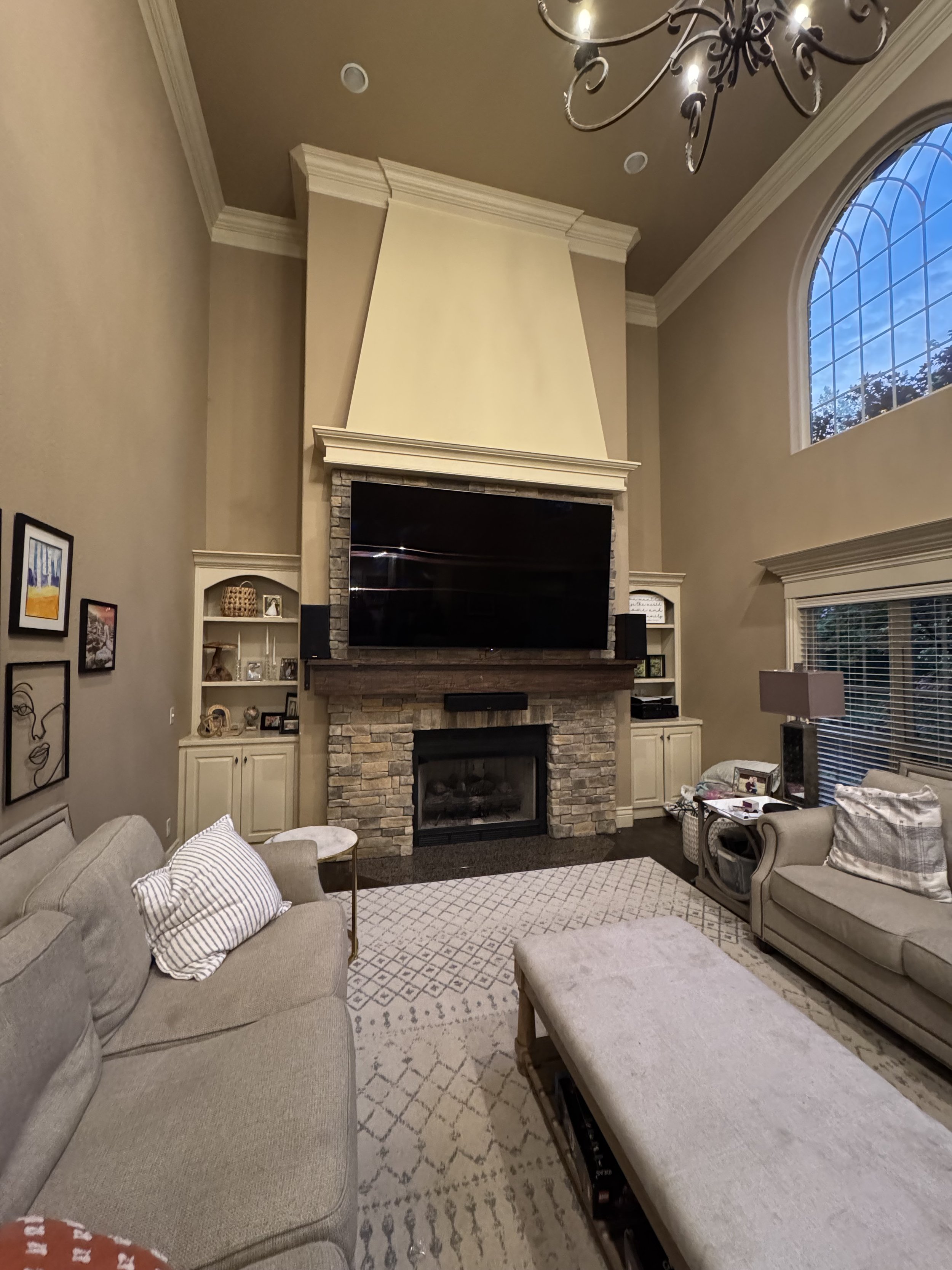 Living room with beige sofas, a fireplace surrounded by stonework, a large flat-screen TV above the fireplace, built-in cabinets on each side, a patterned area rug, and a high ceiling with a chandelier. There are windows showing an evening sky.
