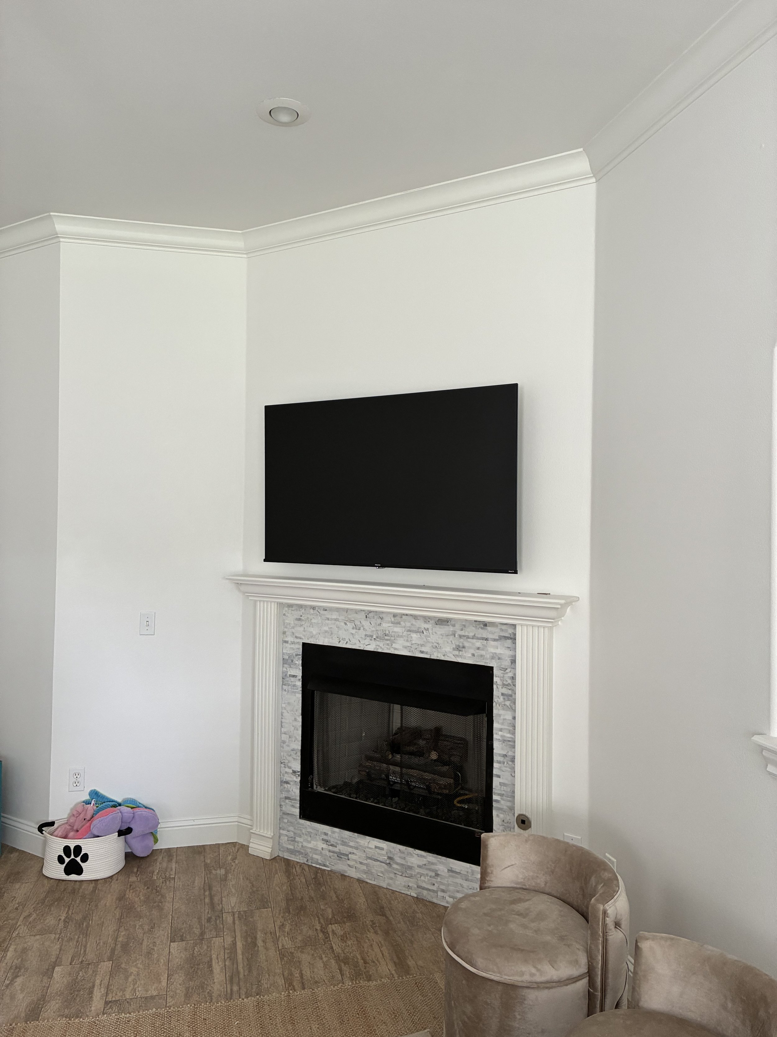 Living room corner with a mounted flat-screen TV above a fireplace surrounded by white trim and gray stone, beige velvet armchairs, and a basket of plush toys on the floor.