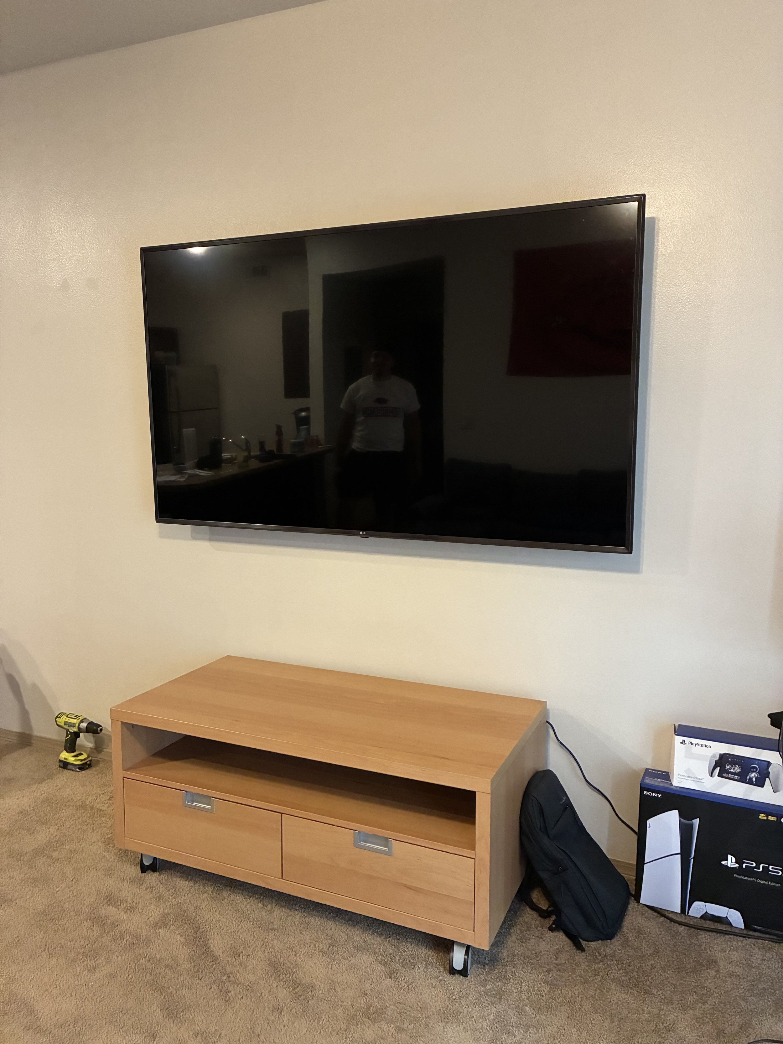 Living room with a flat-screen TV mounted on a beige wall, a wooden TV stand with wheels below it, a black backpack on the floor next to a box of PlayStation 5, and a yellow power drill on the carpeted floor.