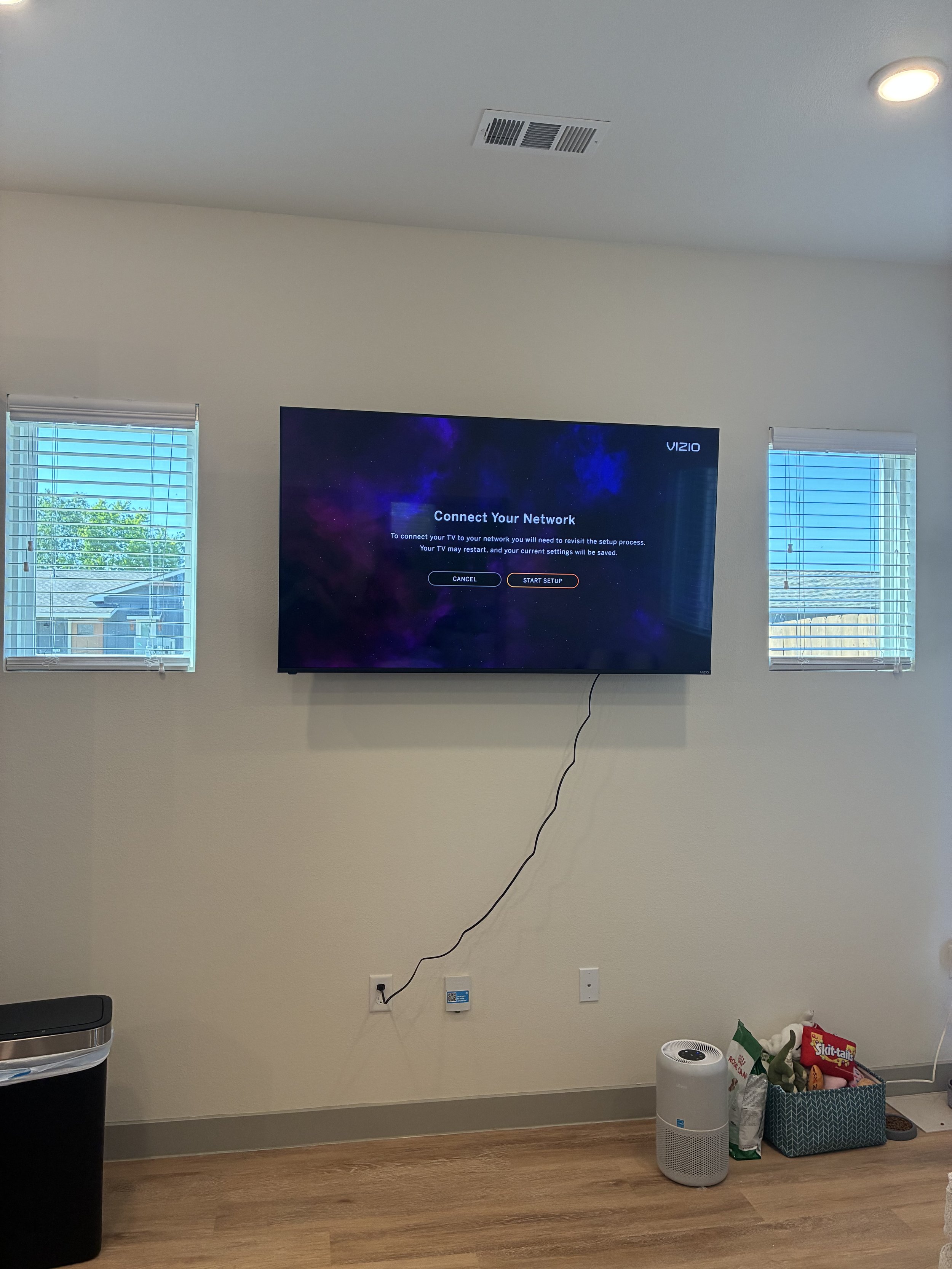 A flat-screen TV on a wall displaying a network connection setup screen, with two small windows to the left and right, a small air purifier and a box of snacks on the floor to the right, and a trash can to the left on a wooden floor.