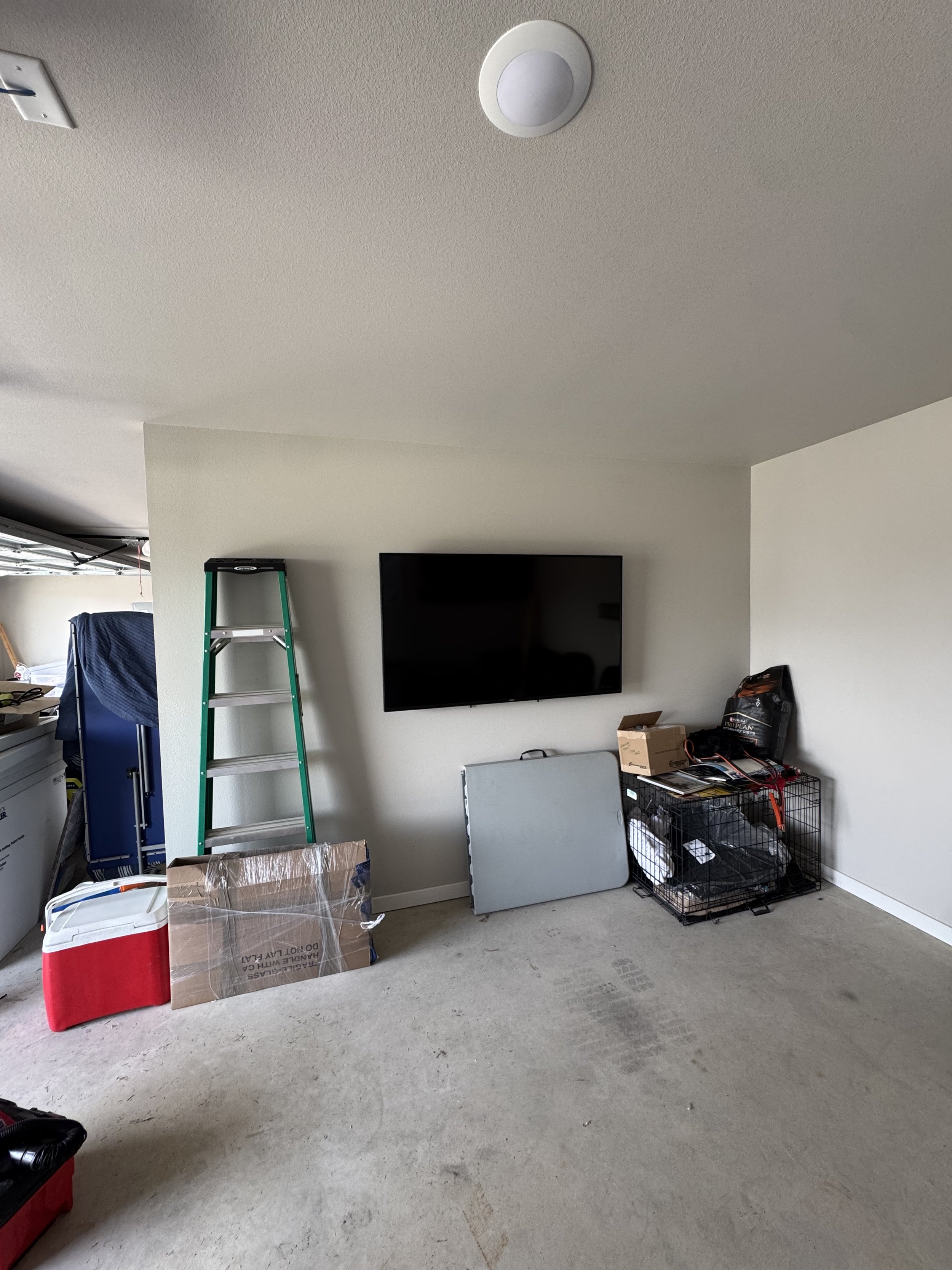 A mostly empty room with beige carpet and white walls. A flat-screen TV is mounted on the wall. There is a green ladder leaning against the wall, a gray folding table, a black wire storage cube with various items, a small cardboard box, a black bag, 