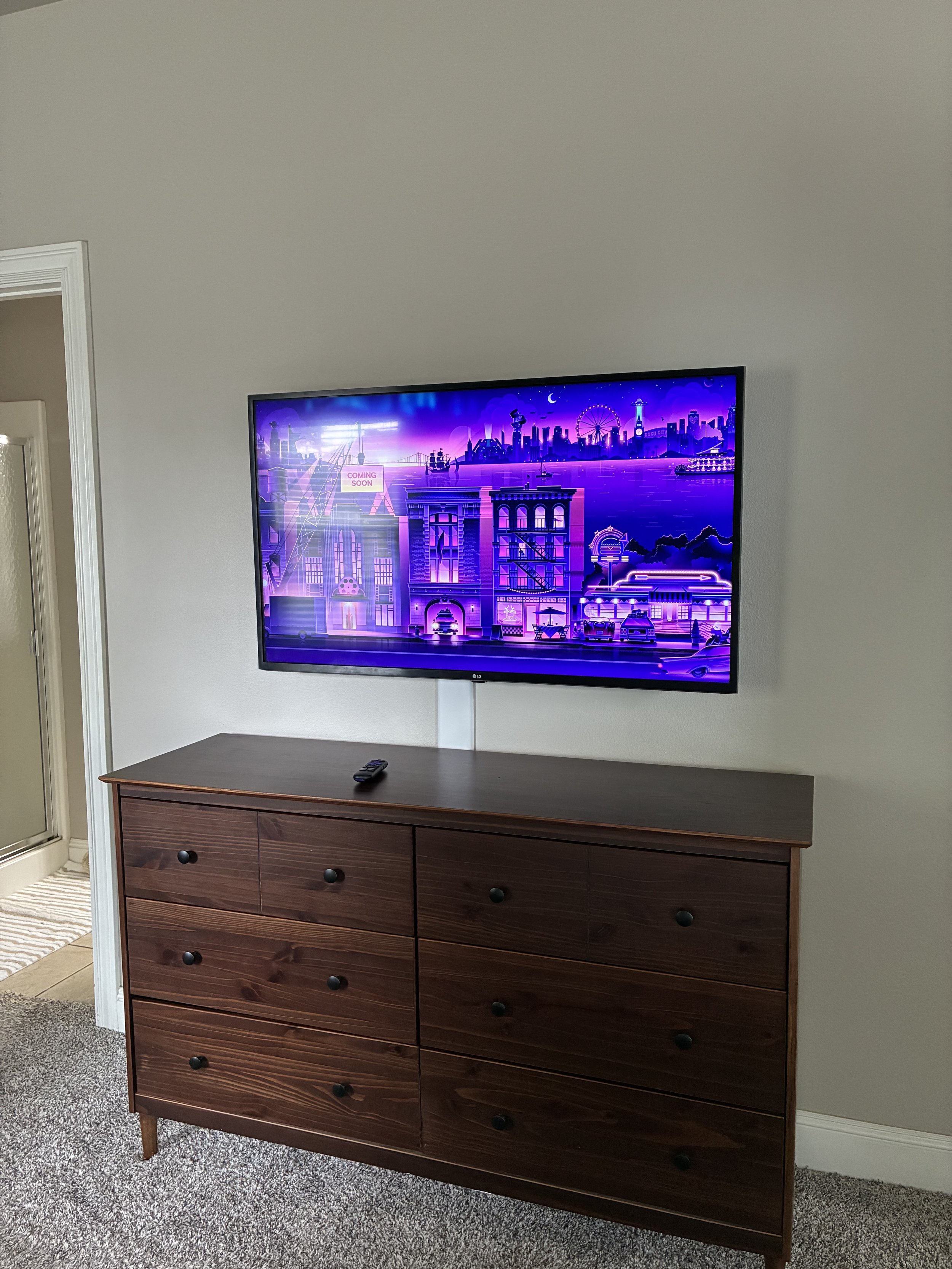 A flat-screen TV mounted on a gray wall above a dark wooden dresser with six drawers, with a remote control on top, in a room with a beige carpet.