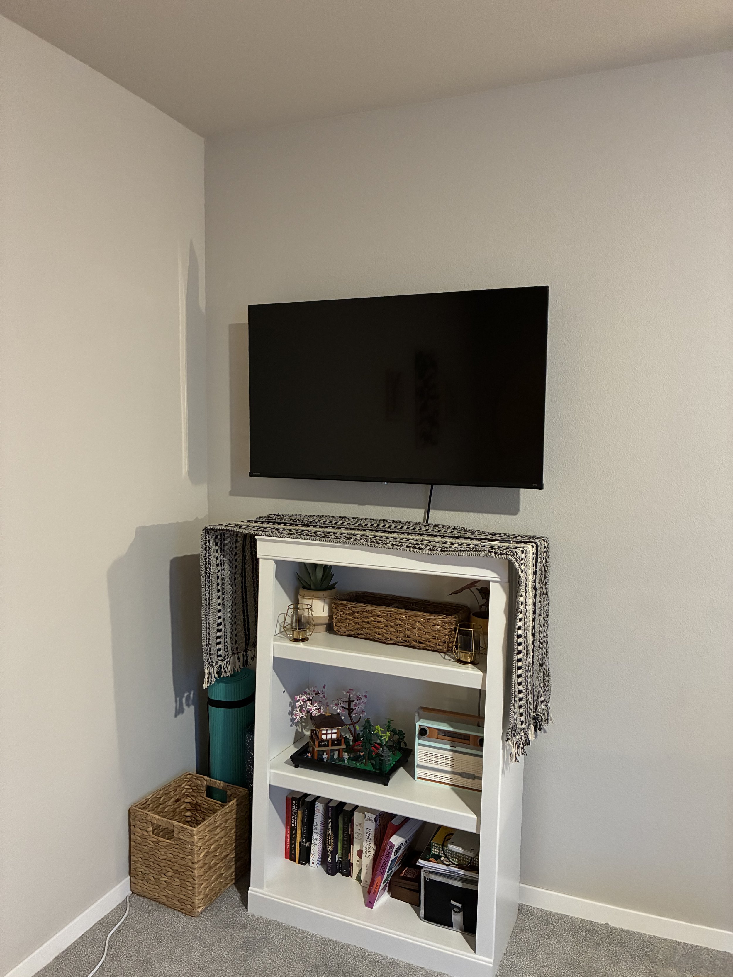 TV mounted on wall above a white bookshelf decorated with a striped throw, small plants, a wicker basket, books, and decorative items in a cozy living room.
