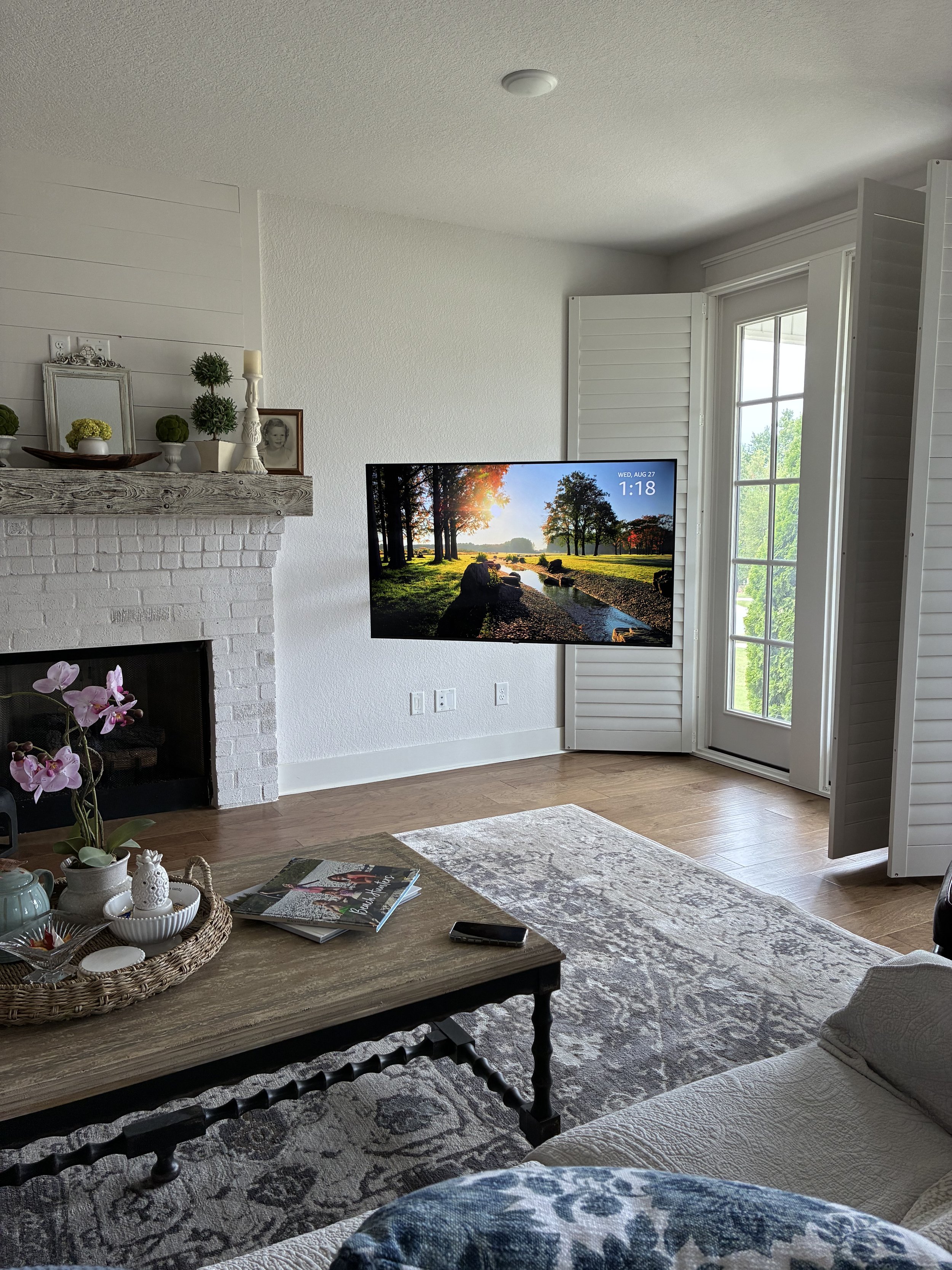 Living room with a white brick fireplace, a flat-screen TV showing a nature scene, a coffee table with magazines and a orchid plant, and open white shutters on a glass door letting in natural light.