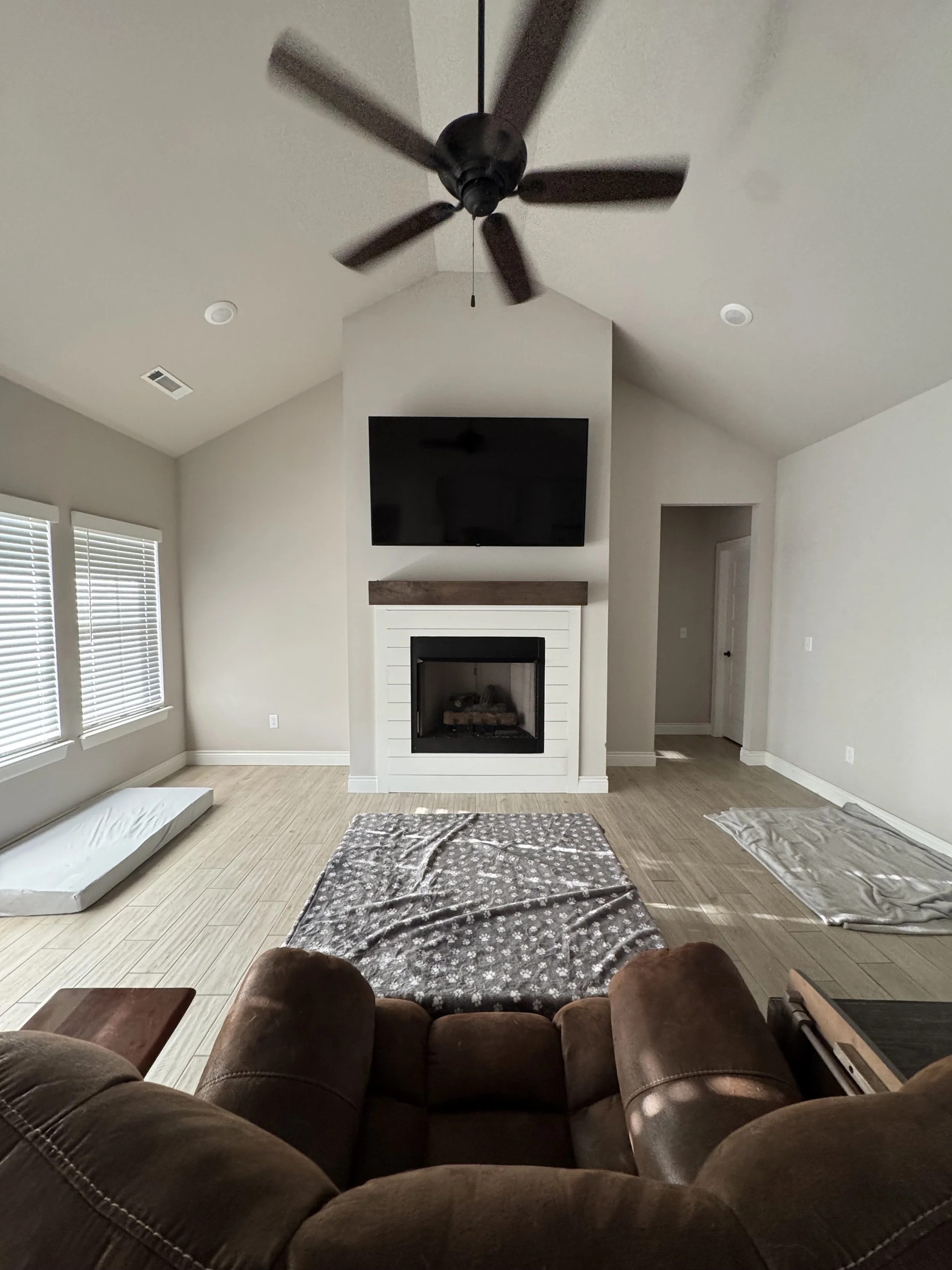Living room with a brown recliner, area rug, fireplace with a mounted flat-screen TV above, three windows with blinds, a ceiling fan, and some blankets on the floor, with light-colored walls and wood-look flooring.