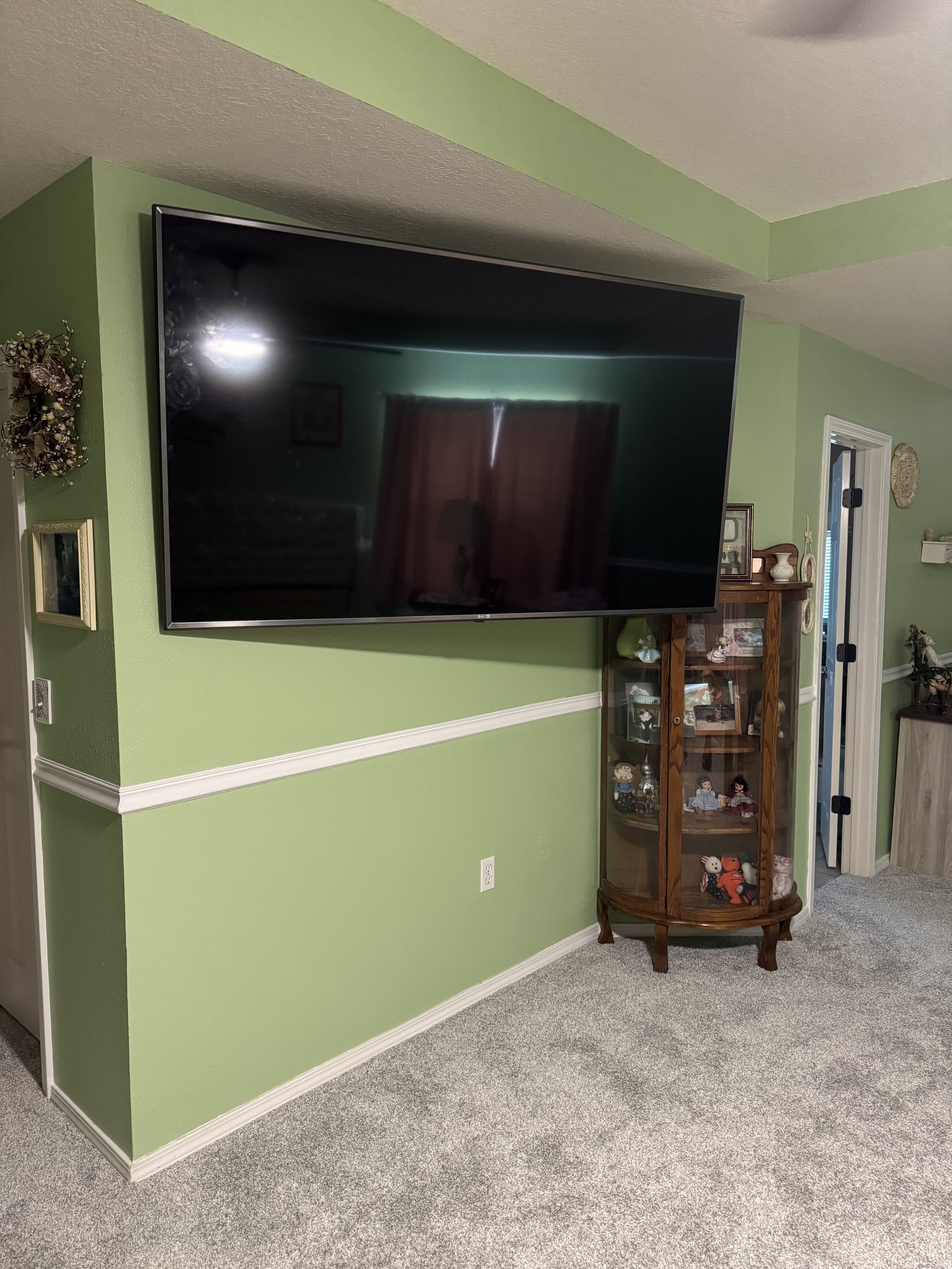 A living room with a large flat-screen TV mounted on a light green wall, a wooden display cabinet filled with decorative items, and a doorway leading to another room.