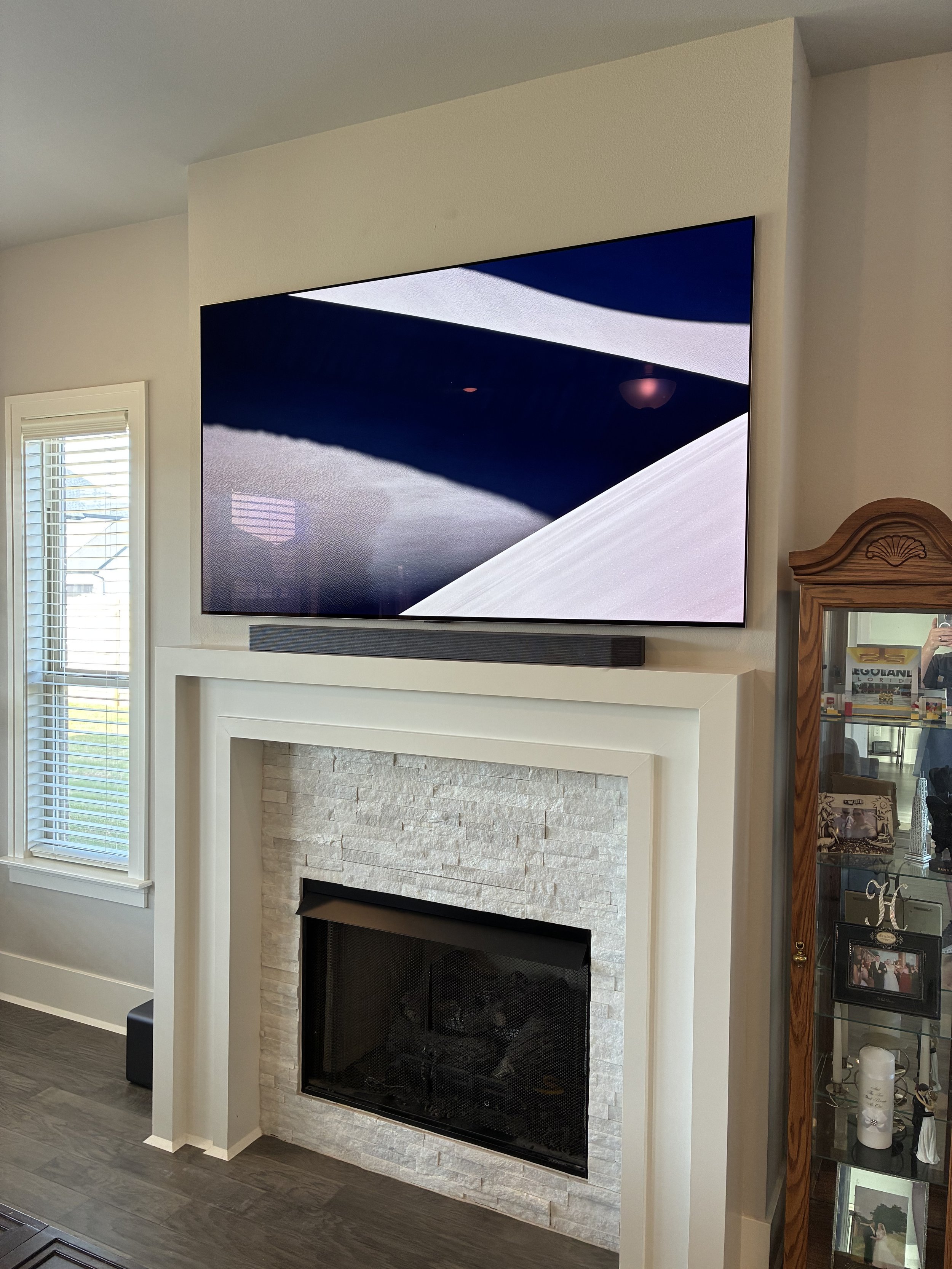 Living room with mounted flat-screen TV above a white stone fireplace with a black metal screen, and a glass display cabinet with family photos and decorative items on the right.