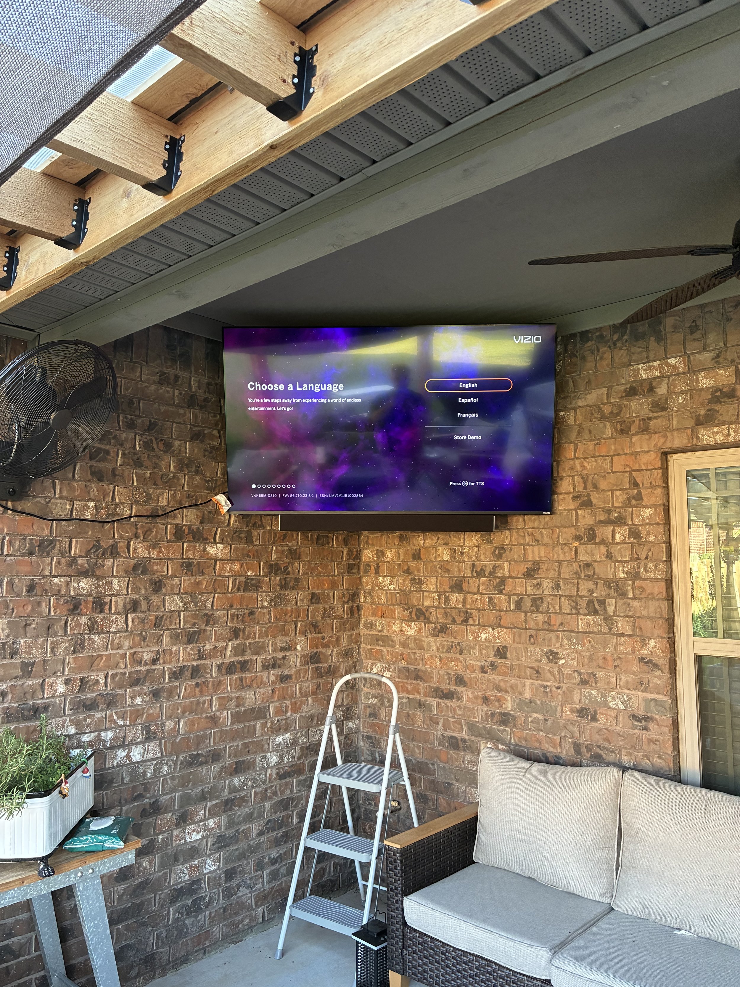 A wall-mounted TV displaying a 'Choose a Language' menu with options in English, Español, Français, and Store Demo, in an outdoor patio area with brick walls, a ceiling fan, a small white folding ladder, a beige outdoor sofa, a side table, and a blac