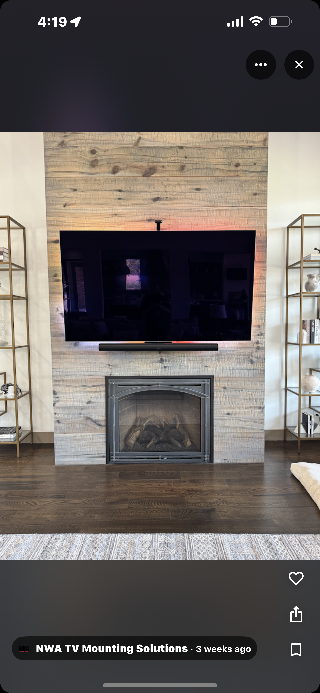 Living room fireplace with a mounted flat-screen TV on a wooden wall, flanked by gold shelving units with decorative items and books, with hardwood floors and a beige rug.