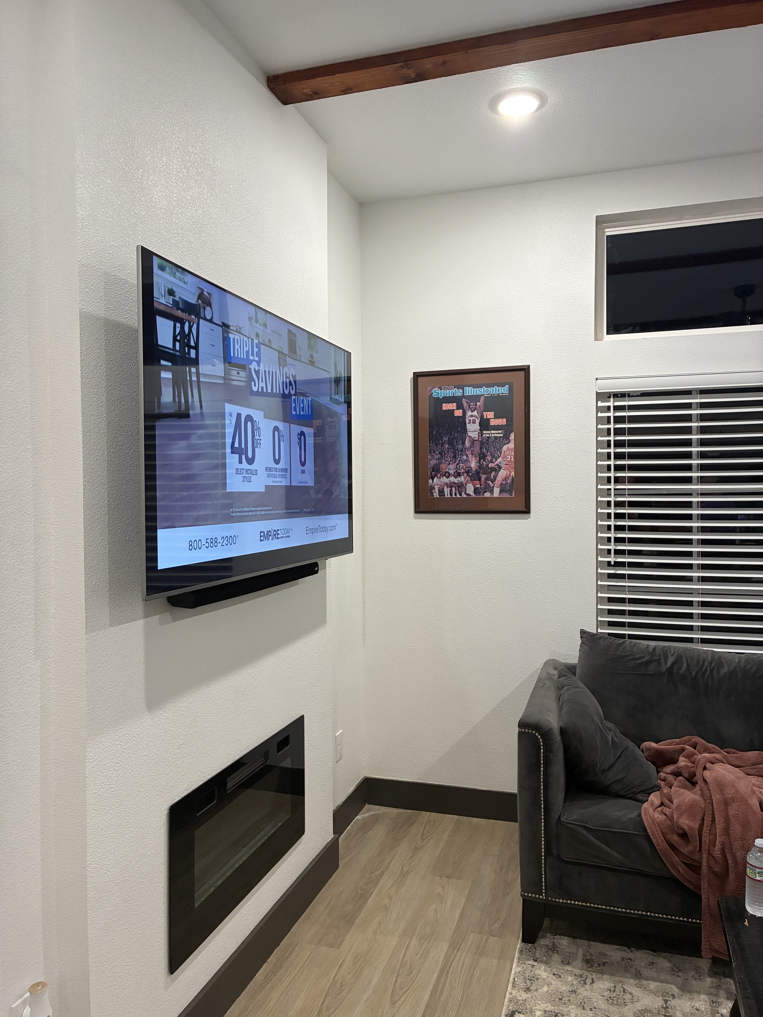 Living room corner with mounted flat-screen TV displaying an advertisement, framed sports magazine cover on white wall, dark grey couch with an orange blanket, window with horizontal blinds, wooden flooring, and ceiling light.