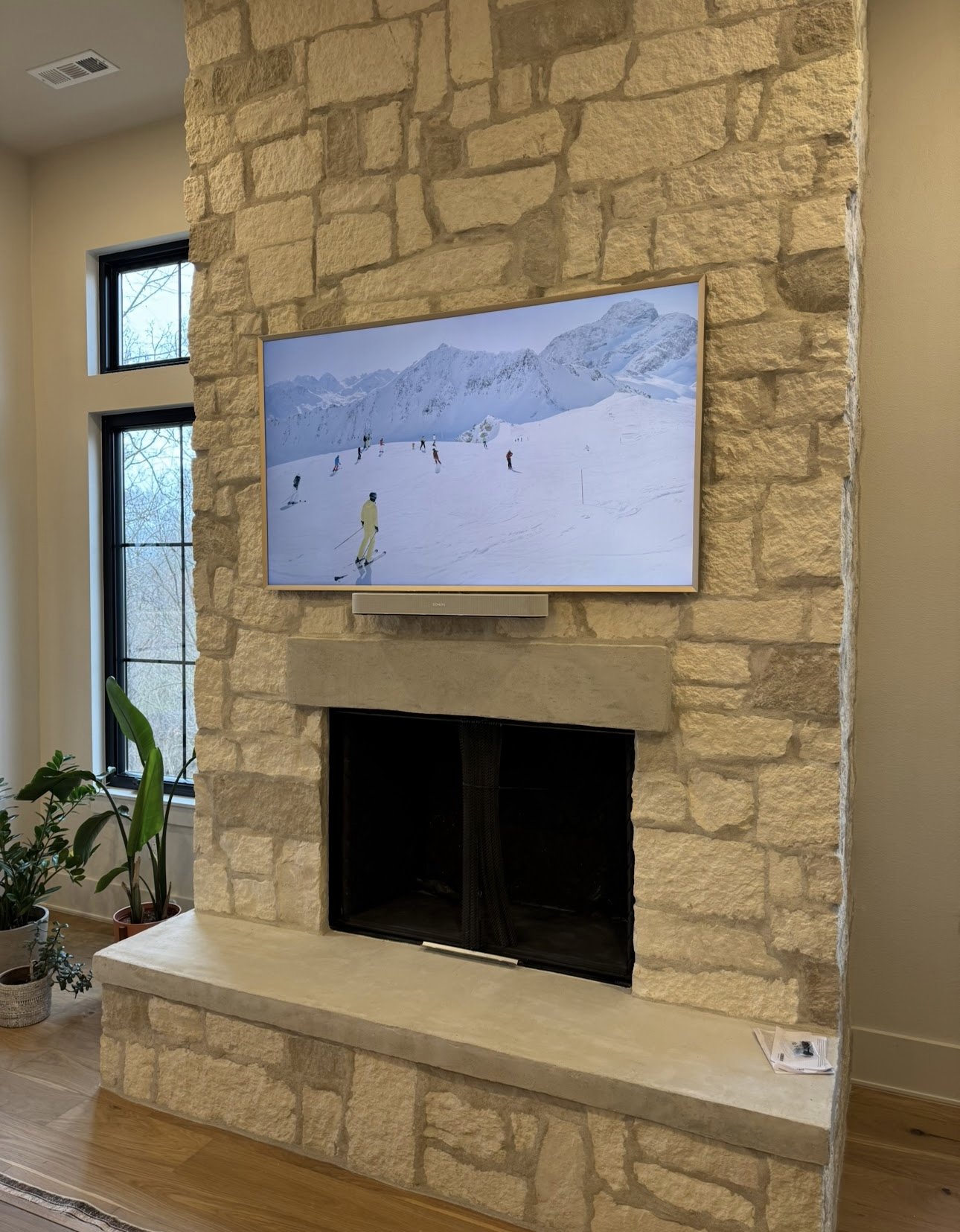 Living room fireplace with a mounted flat-screen TV showing a snowy mountain scene with skiers. Potted plants near the window, natural light, light-colored wall and wood flooring.