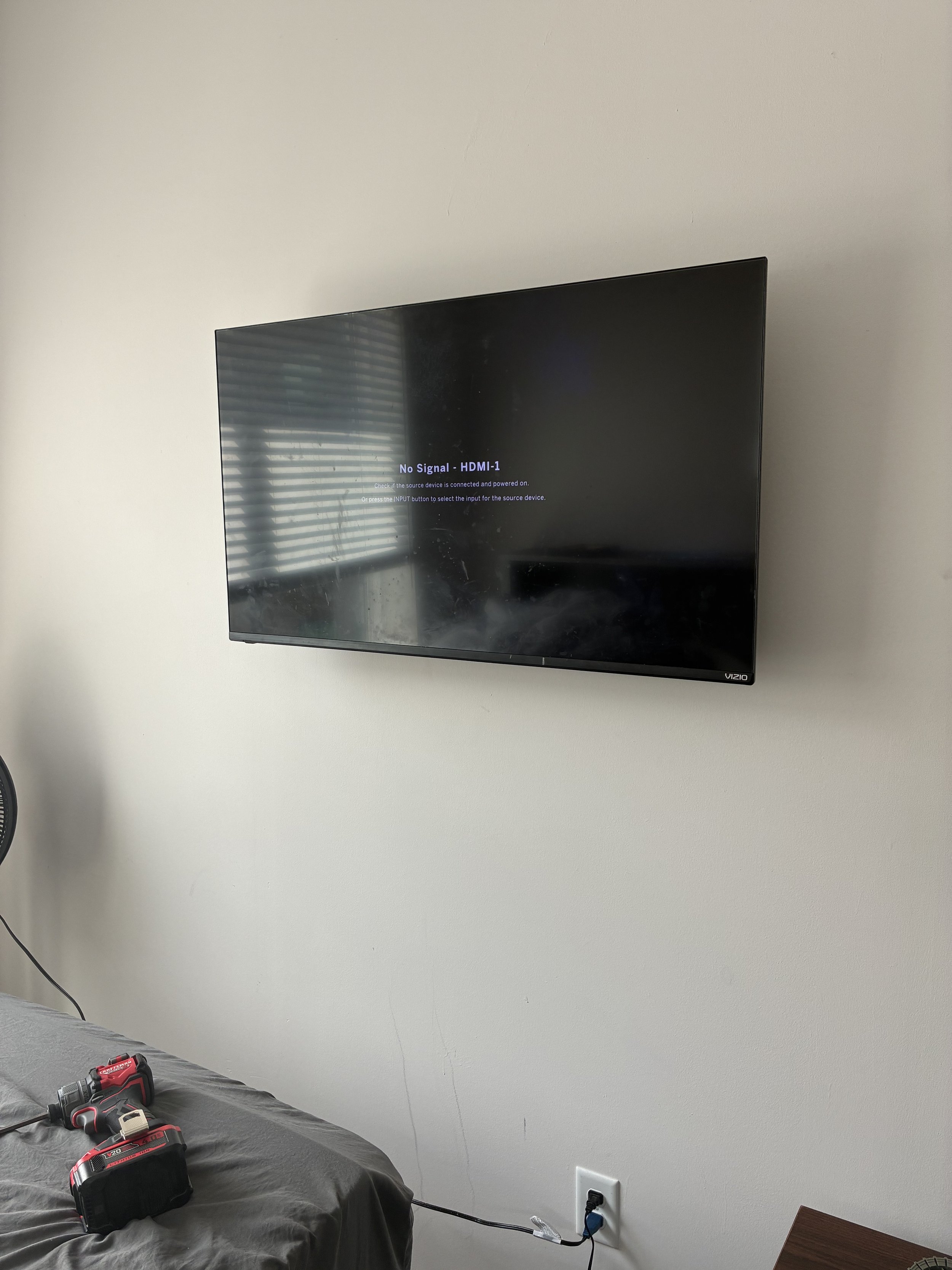 A flat screen Vizio television mounted on a white wall, displaying a "No Signal" message with an HDMI-1 input error. A window with blinds reflected in the screen, and a bed with tools and a power drill nearby are visible.