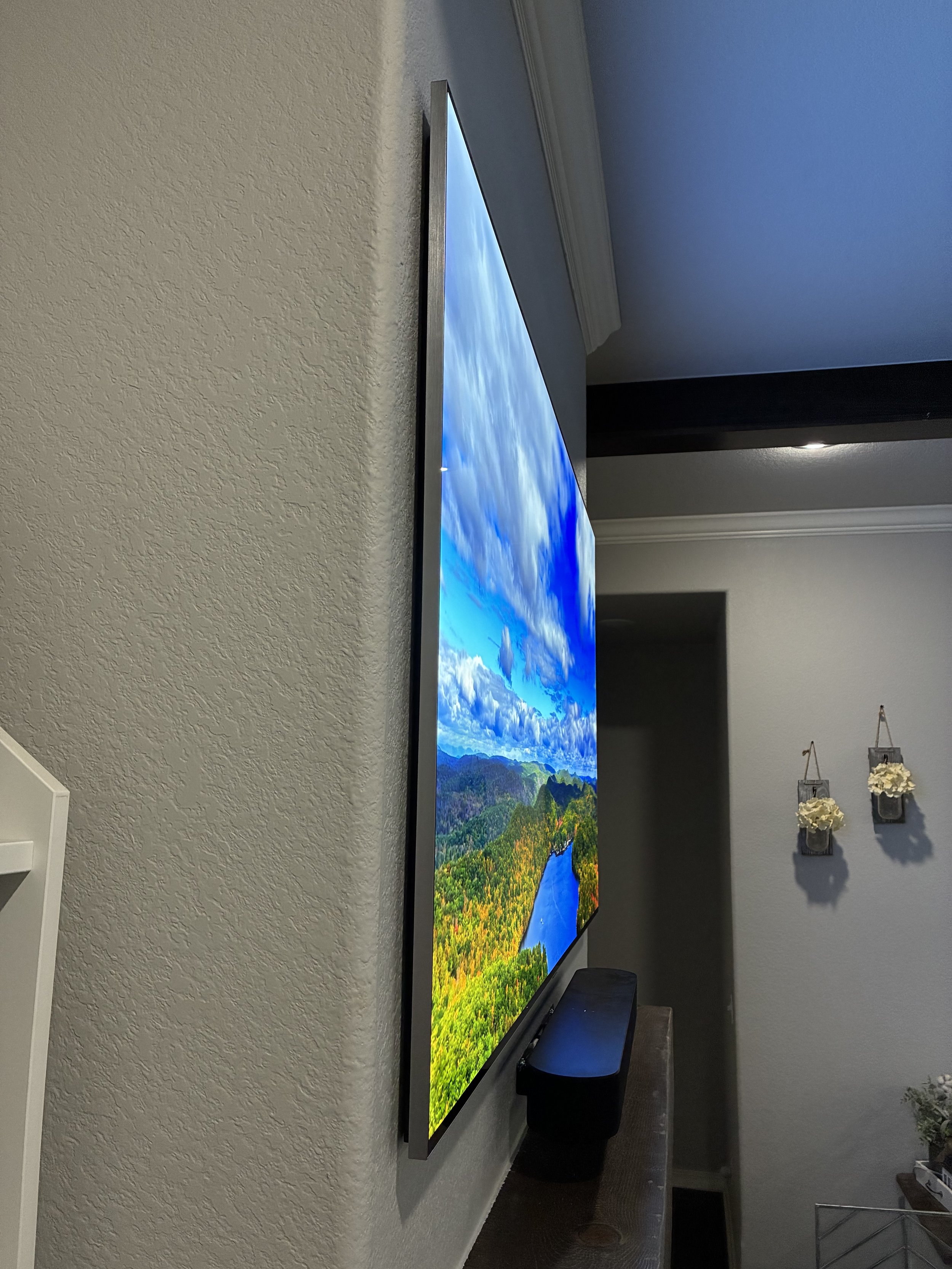 A flat-screen TV mounted on a beige wall displaying a landscape scene with a river, green hills, and a bright blue sky with clouds.