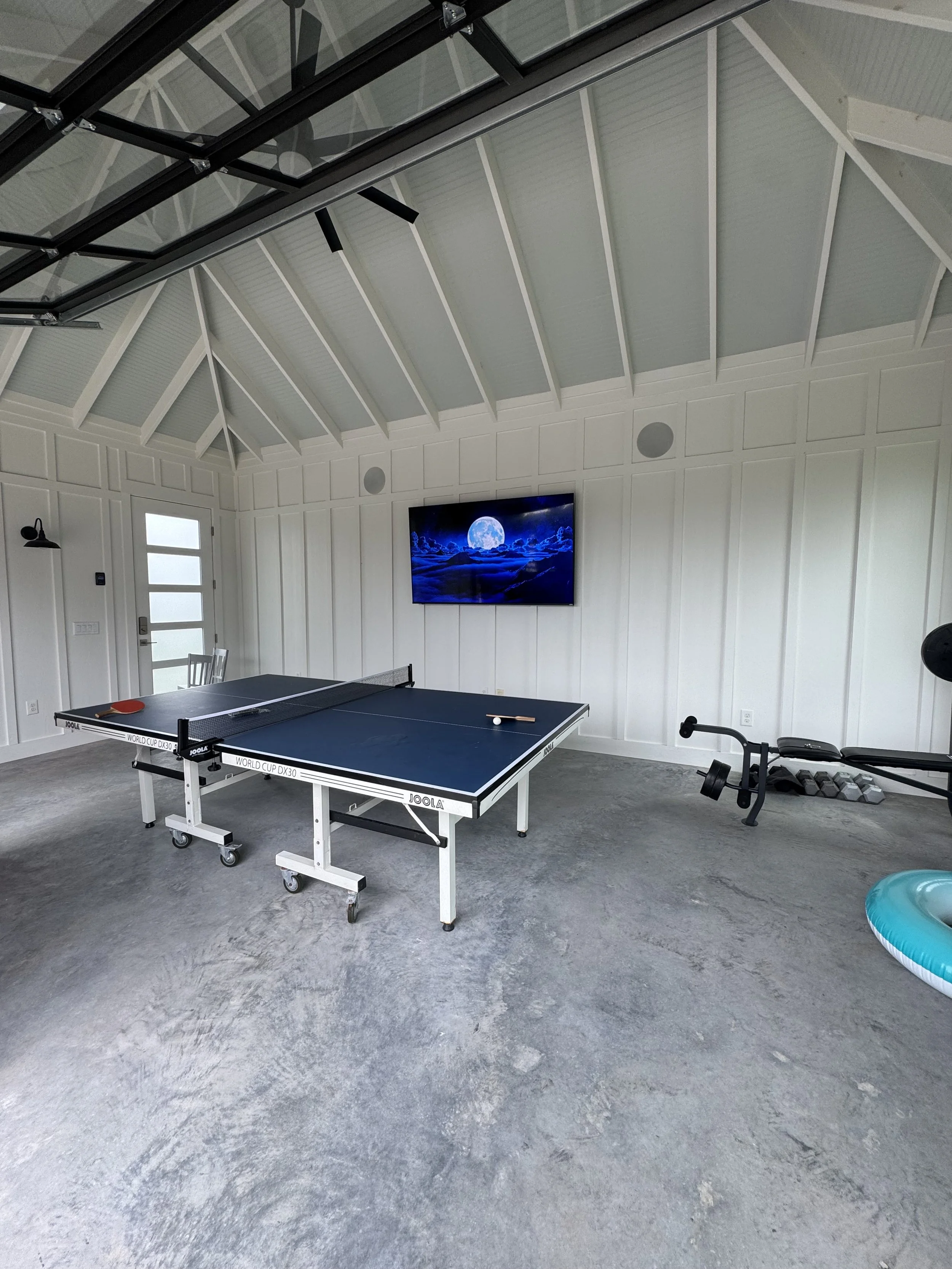 A room with a ping pong table, a wall-mounted TV displaying a moonlit scene, exercise equipment, and a large floathopper pool float in a minimalist style with white paneled walls and a high ceiling.