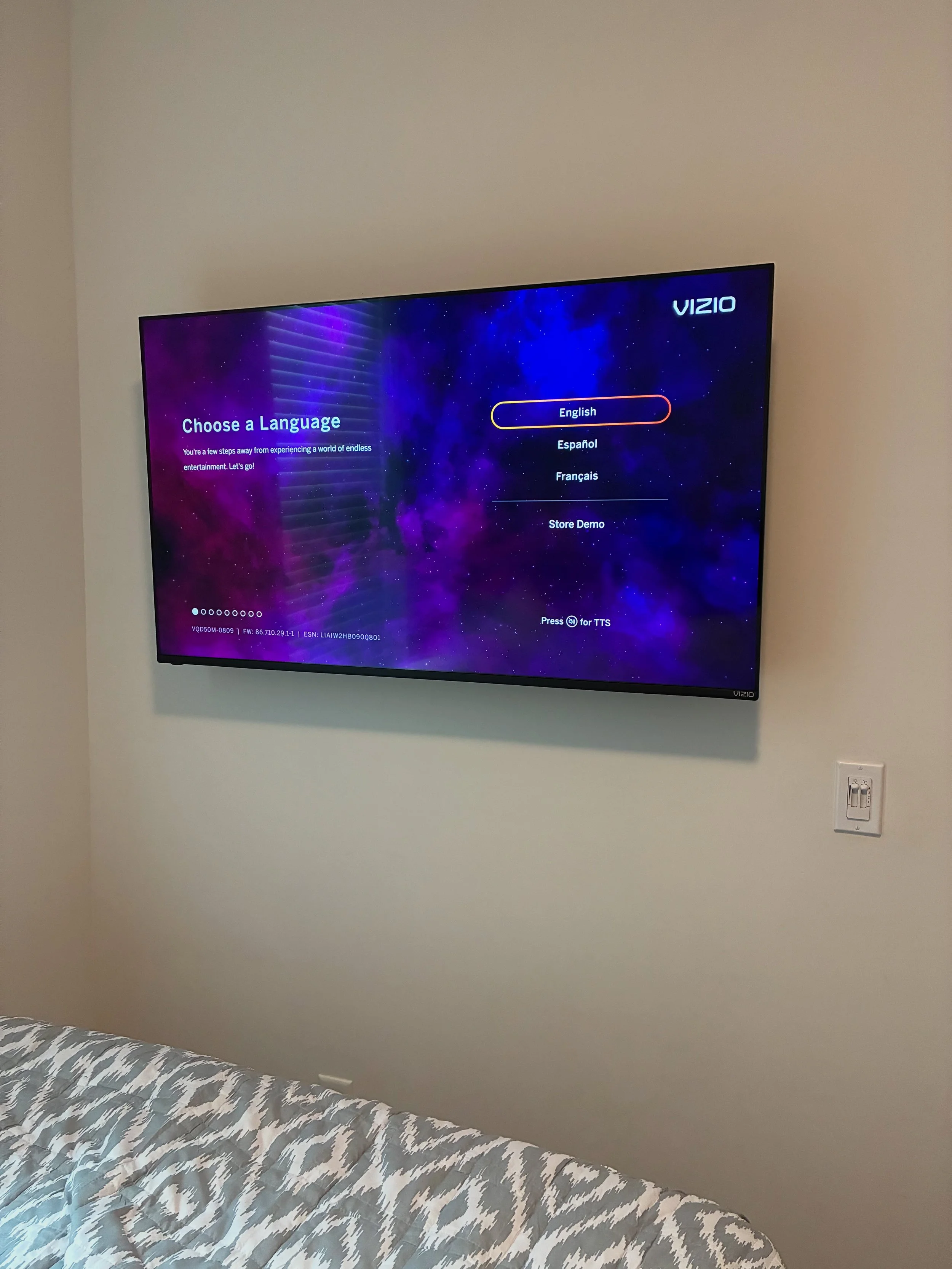 TV screen displaying a language selection menu with options for English, Español, Français, and Store Demo, mounted on a beige wall in a bedroom with a bed with patterned sheets in the foreground.