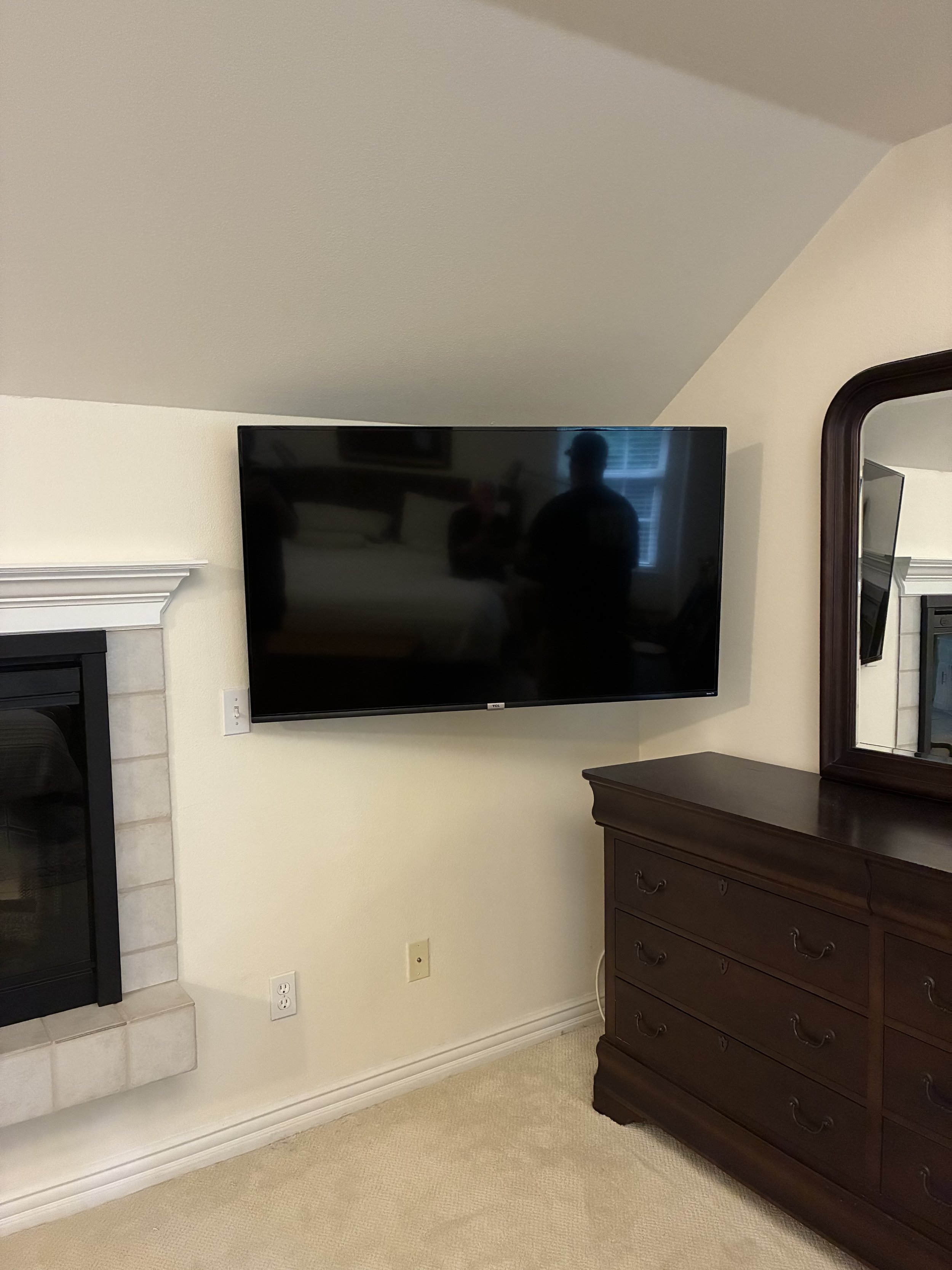 A flat-screen TV mounted on a beige wall above a dark wooden dresser with a mirror in the room.