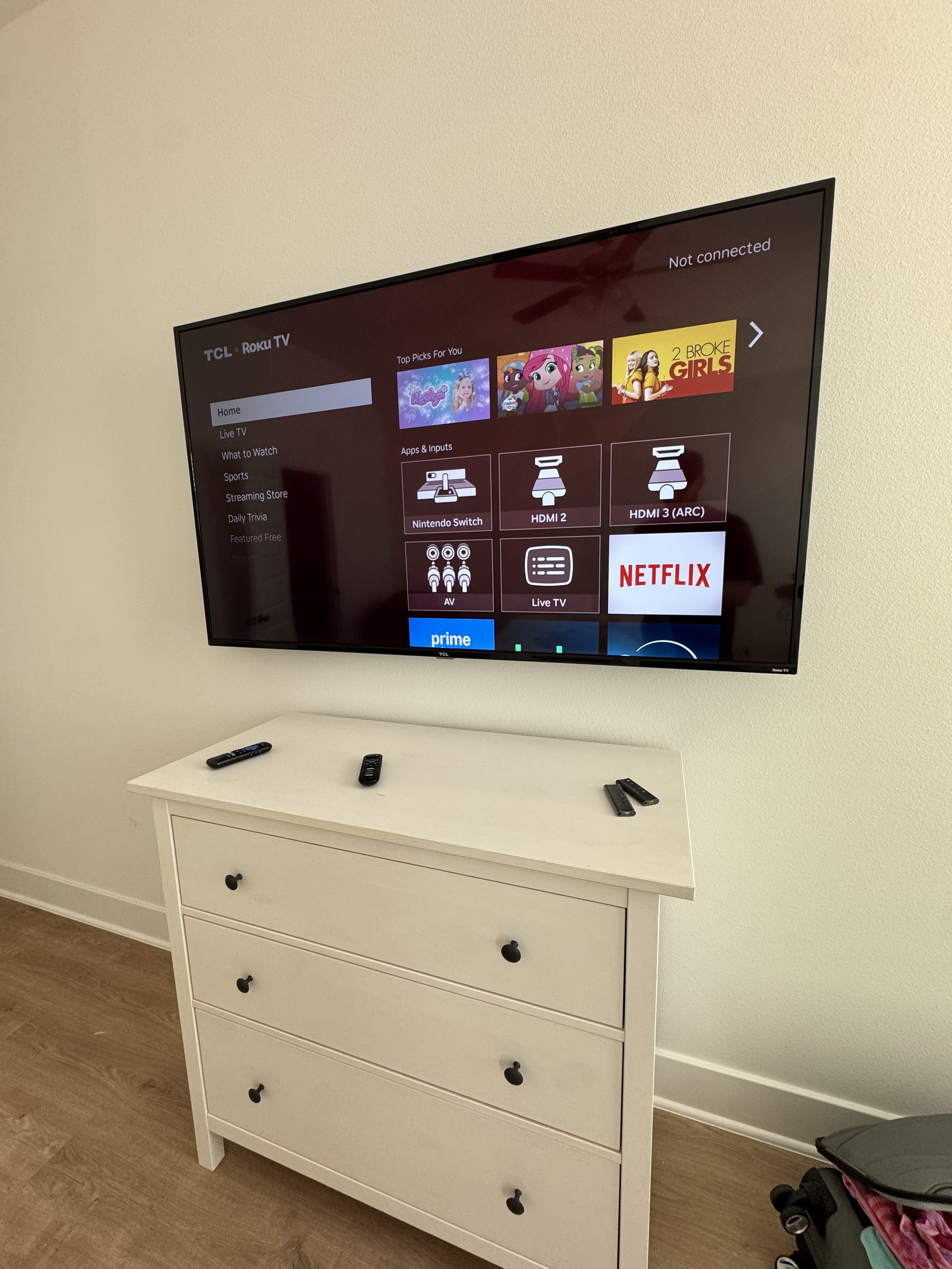 A wall-mounted TCL Roku TV displaying the home screen with options like Live TV, Streaming Store, Daily Trivia, and Netflix. Below the TV, there is a white dresser with three drawers, on top of which are several TV remote controls.