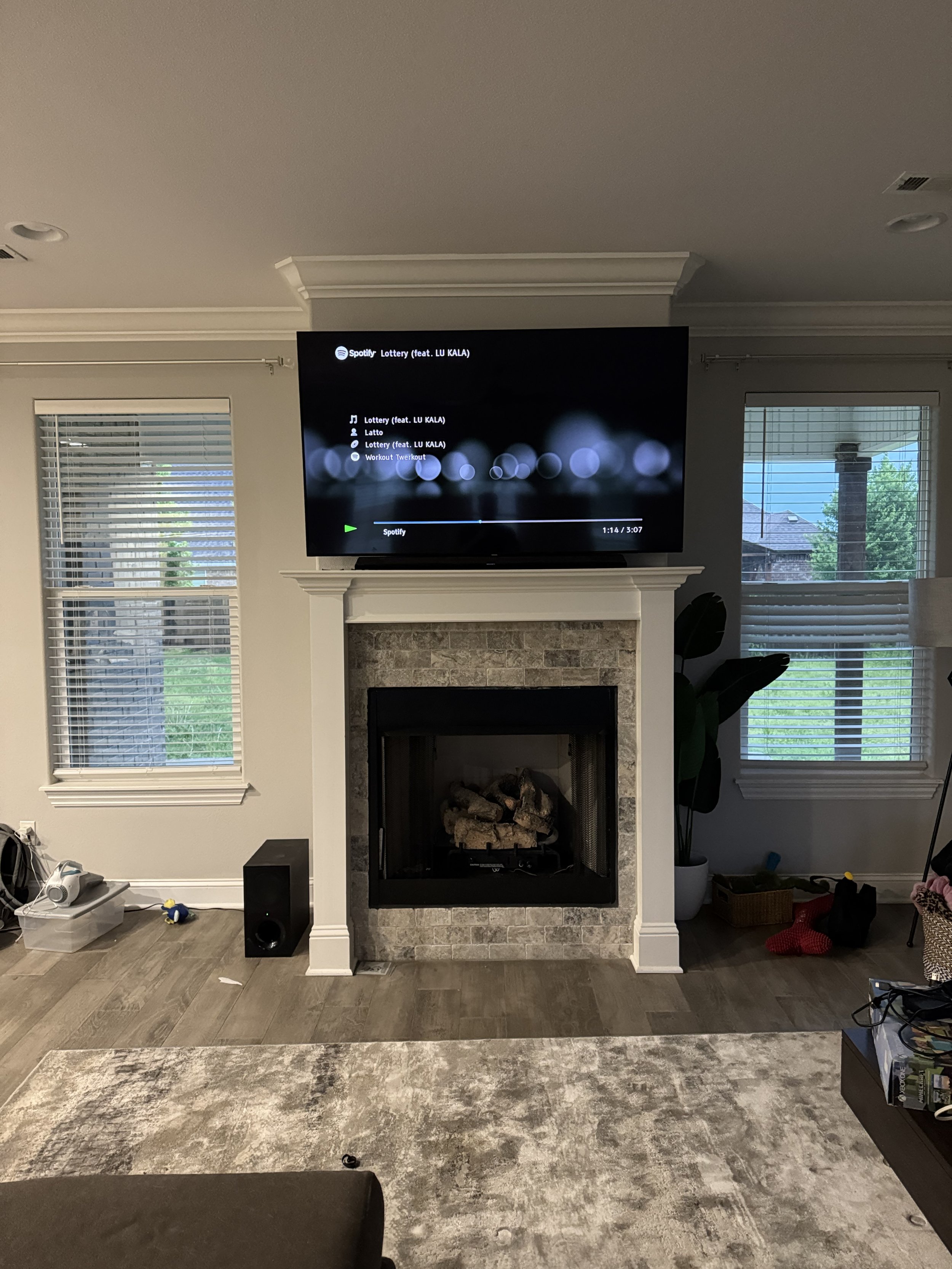 Living room with a fireplace, a mounted TV showing Spotify playlist, two windows with blinds, a black speaker near the fireplace, a box with objects, and a carpeted area with some scattered items.