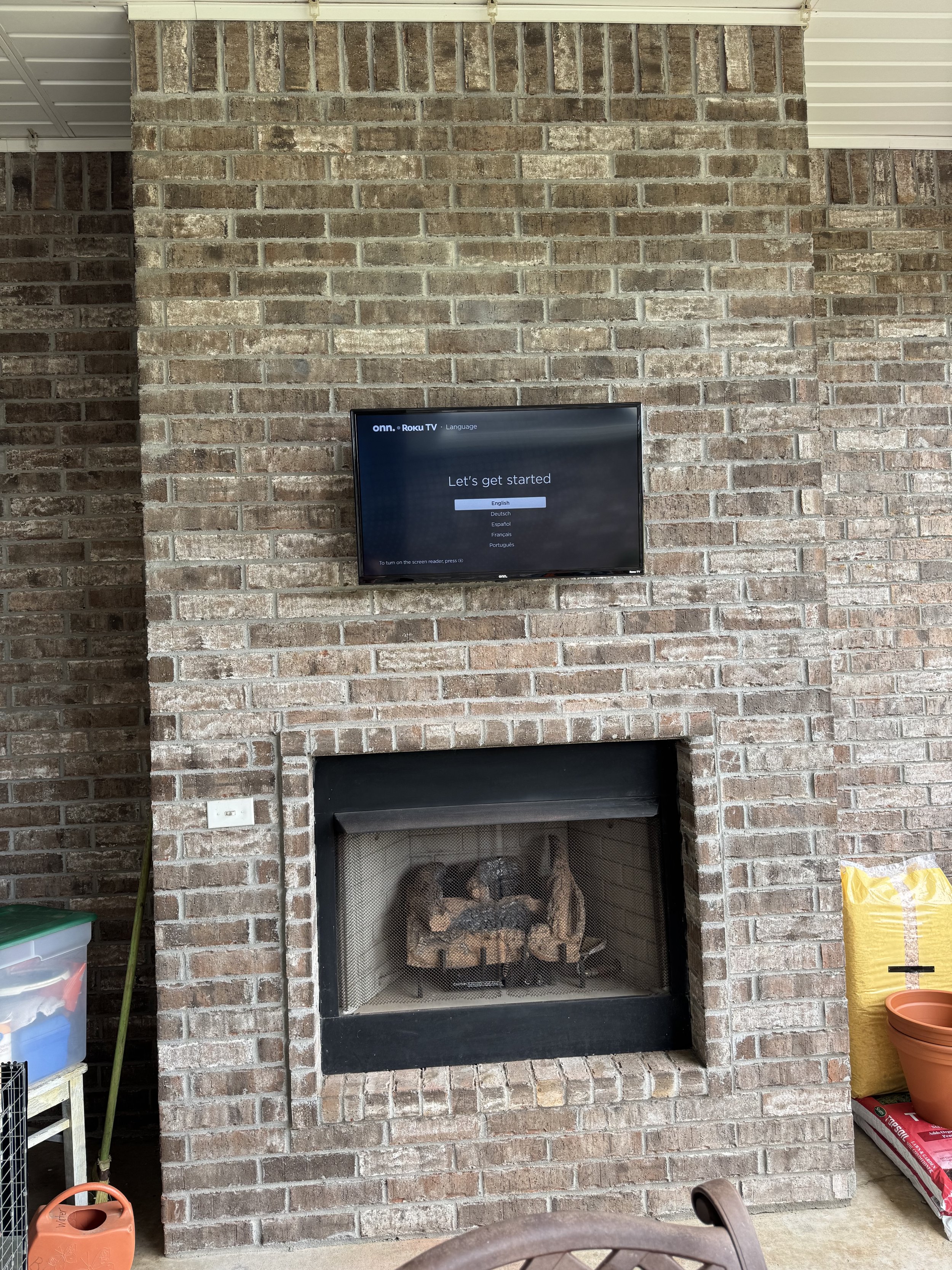 A brick fireplace with a mounted flat-screen TV displaying a setup menu on top of the fireplace. To the left, there's a plastic storage container, and to the right, a yellow bag, a flowerpot, and some gardening supplies on the floor.