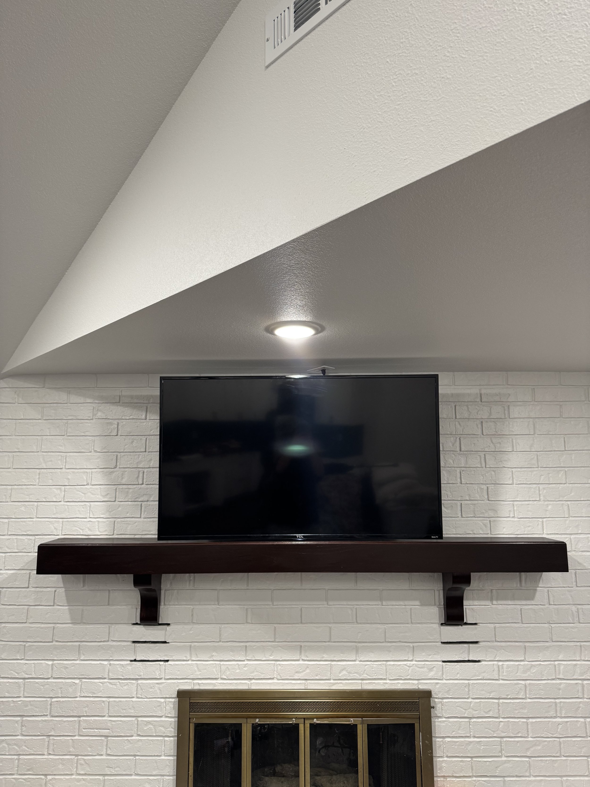 TV mounted above a fireplace on a white brick wall with a dark wooden shelf and a vent in the ceiling.